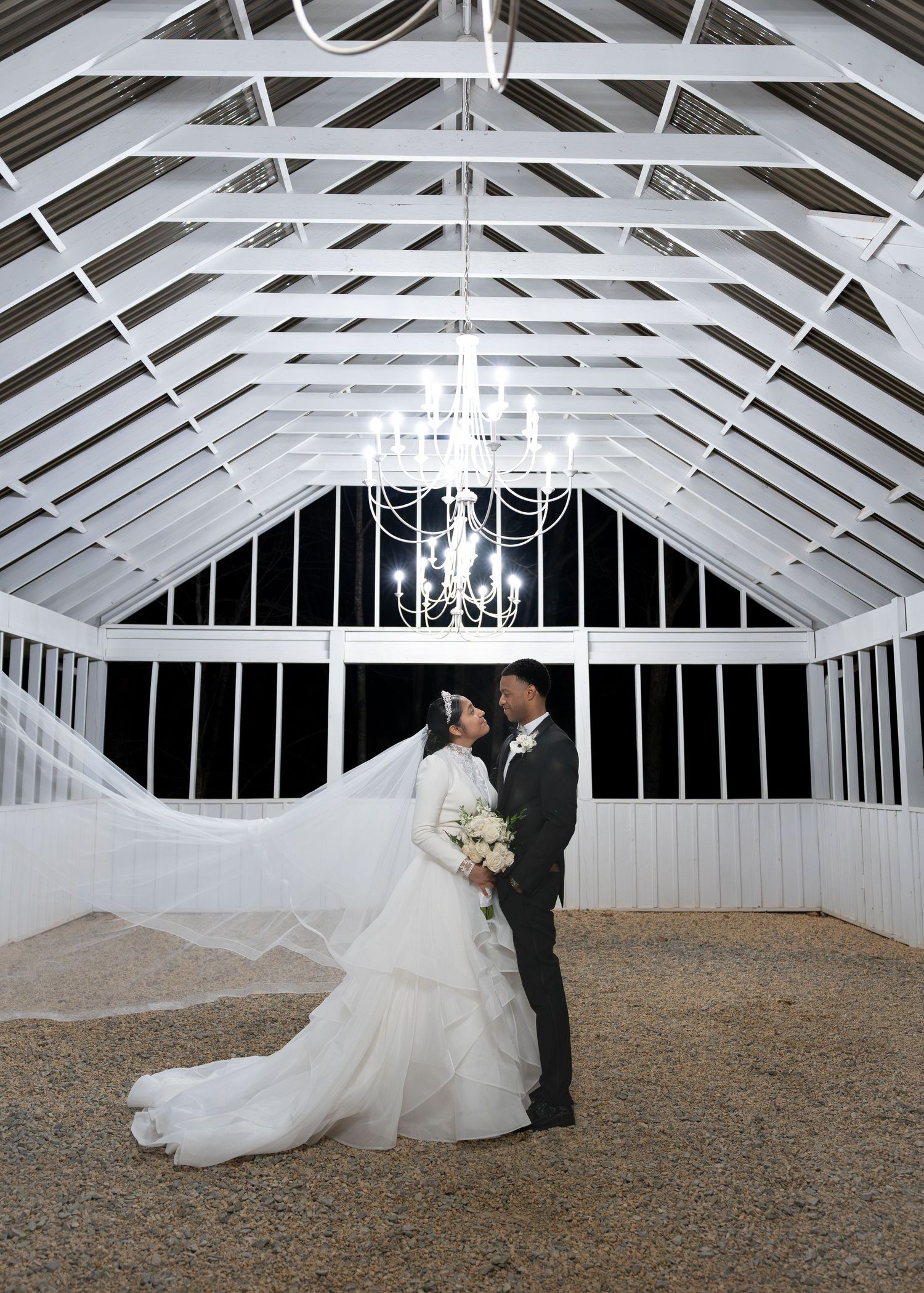 Bride and groom share a romantic moment inside a white barn venue with chandelier overhead.