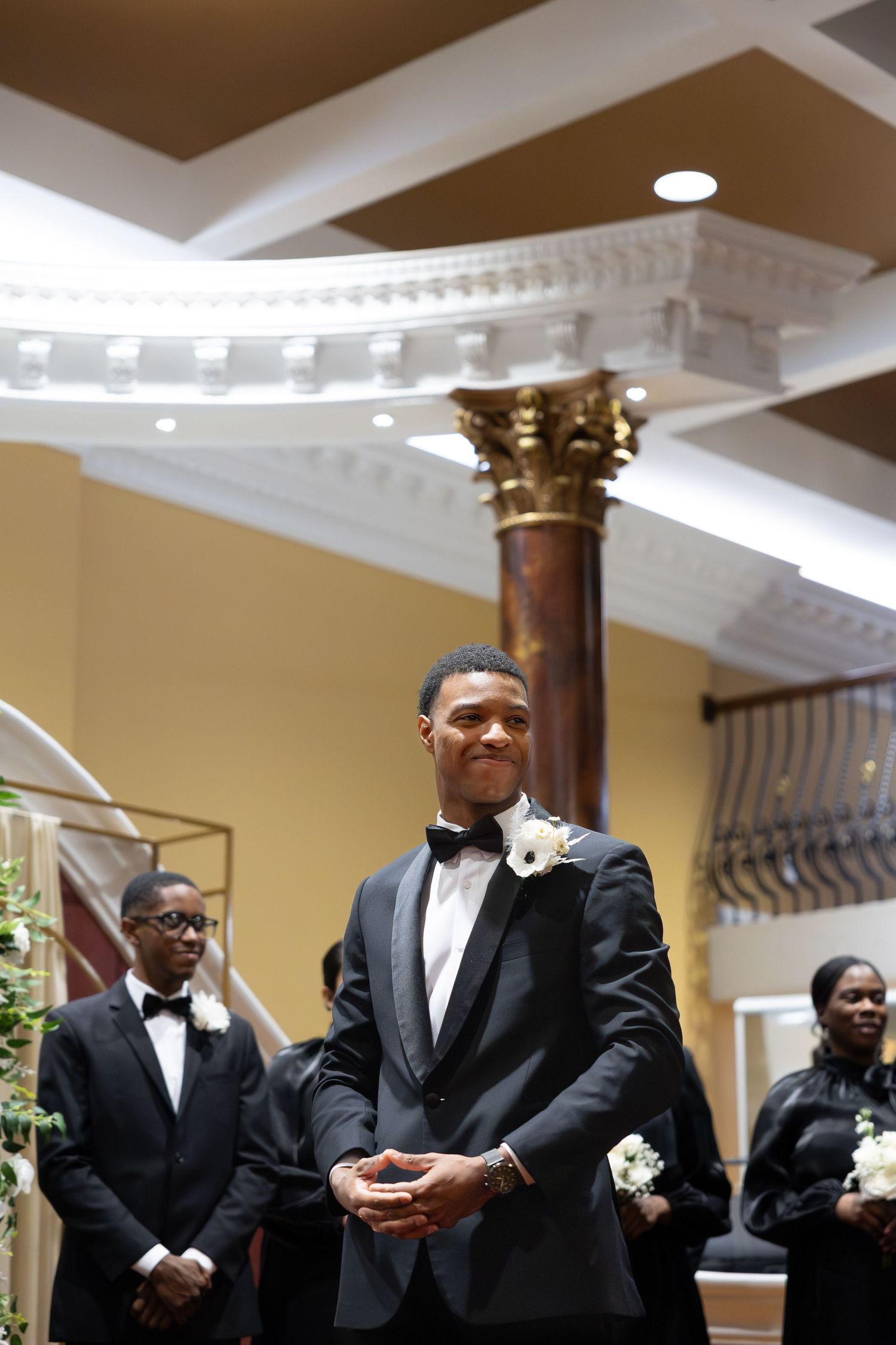 Smiling groom in black tuxedo with white boutonniere awaiting bride at wedding altar.
