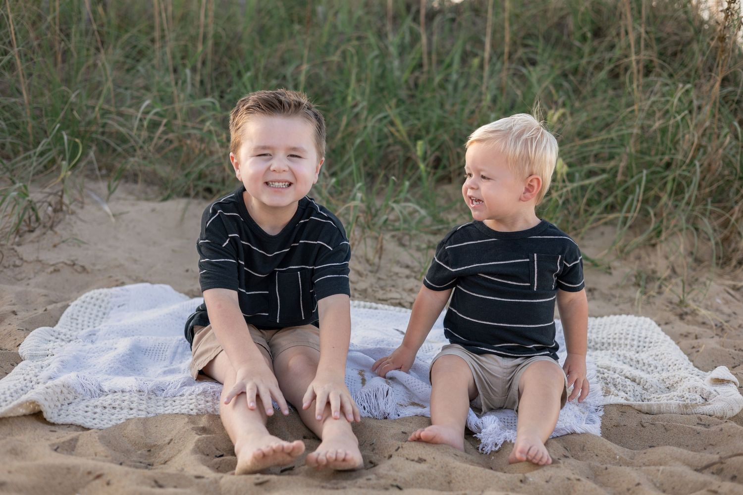 Two young boys in matching navy striped shirts sit smiling on a white blanket on a sandy beach.