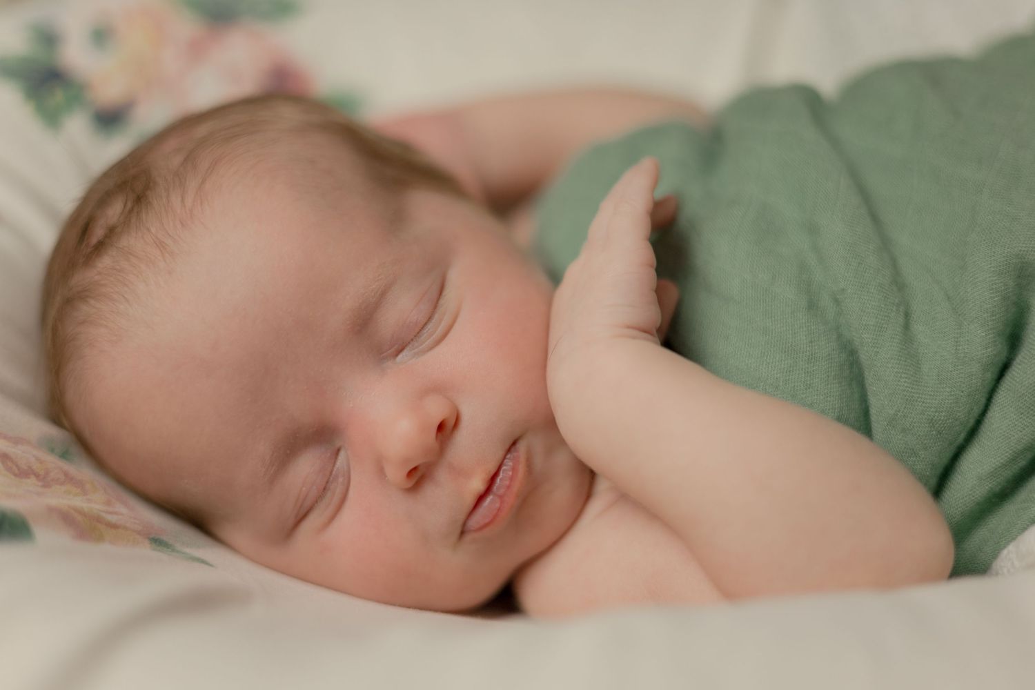 Serene newborn infant sleeps soundly wrapped in green muslin swaddle on white floral surface.