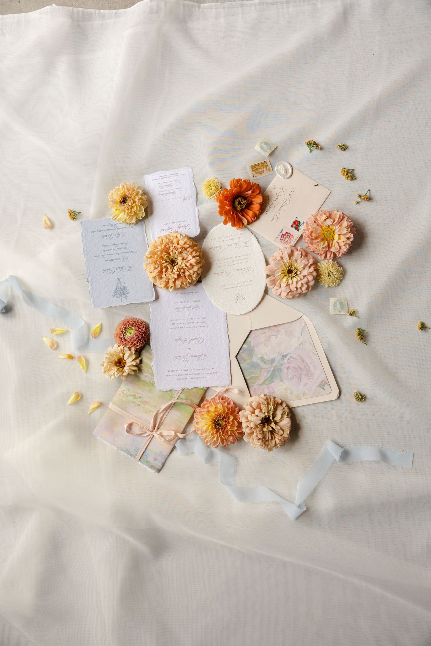 Colorful wedding stationery flat lay surrounded by orange and yellow dahlias on white fabric.