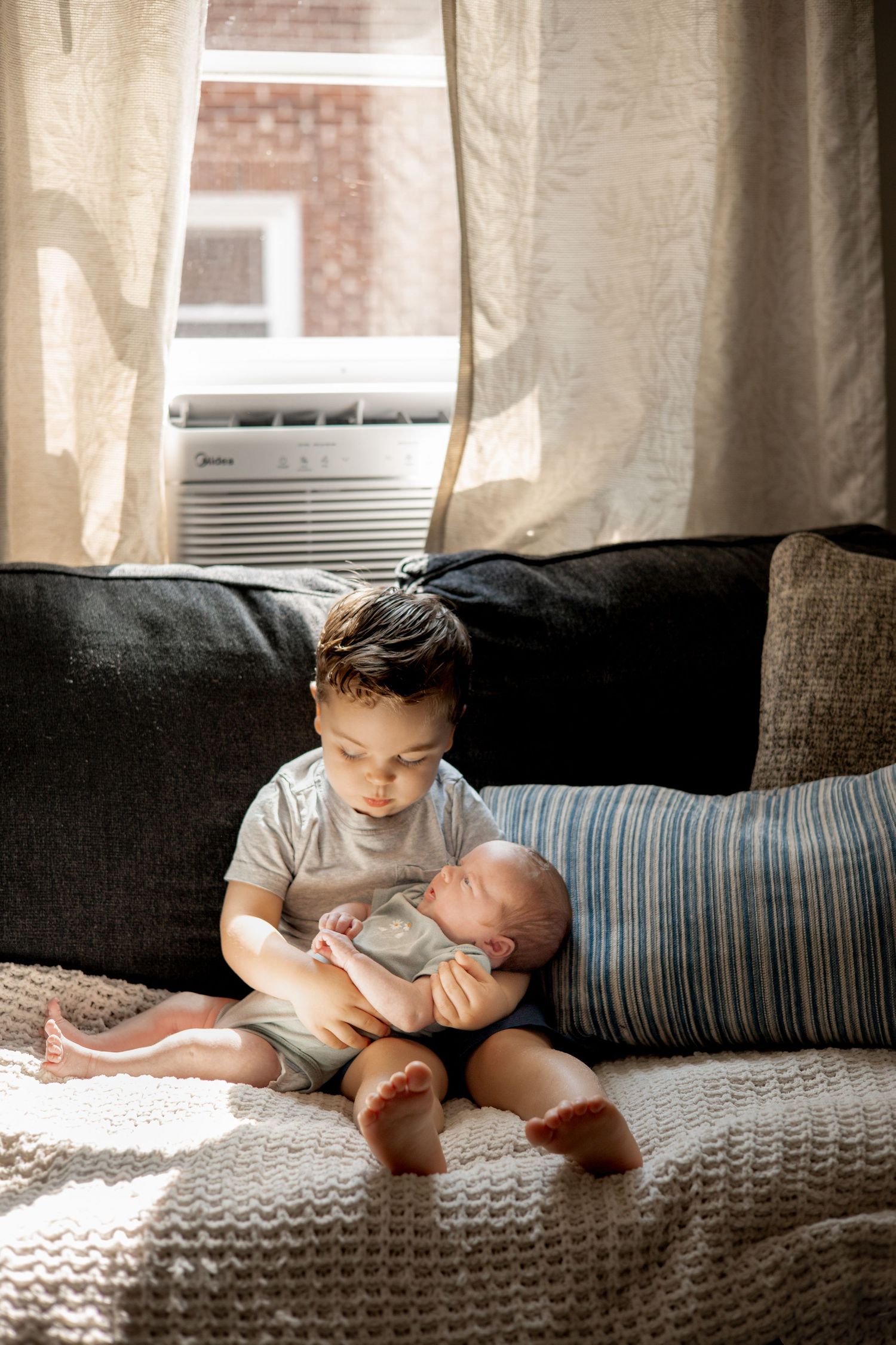 Young boy gently holds sleeping newborn baby sibling on a couch by a sunny window.