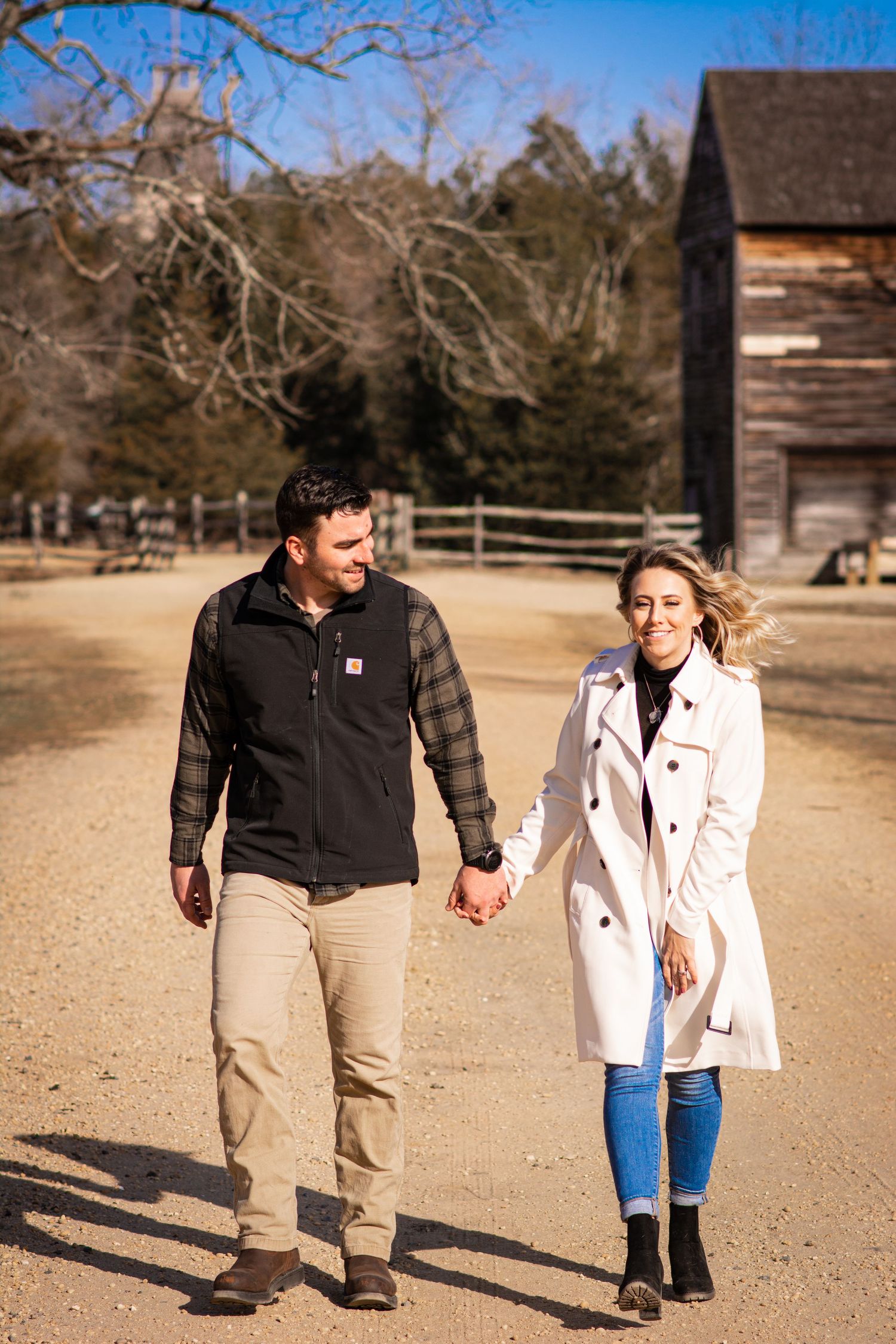 Happy couple holding hands walking down a sunny dirt path near a rustic wooden barn in winter.