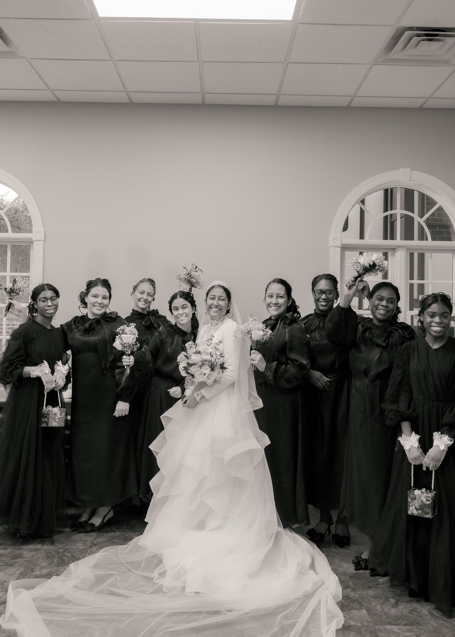 Black and white photo of bride in white gown with bridesmaids in dark dresses at wedding venue.
