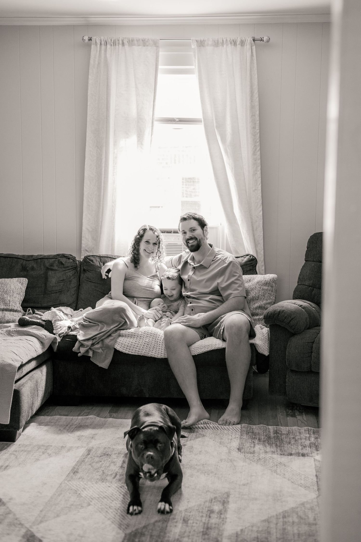 Black and white family portrait on couch with dog in foreground, parents and child smiling by window.