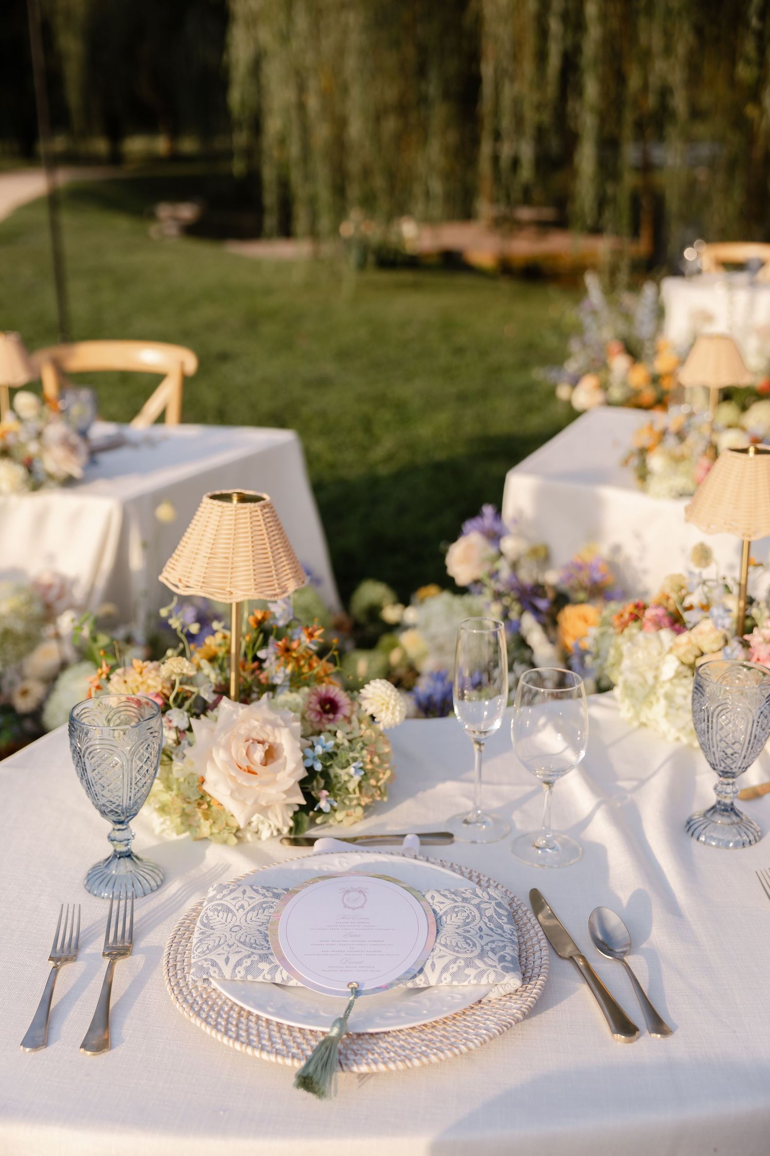 Elegant outdoor wedding place setting with blue glassware, floral centerpiece, and rattan lamp on white tablecloth.