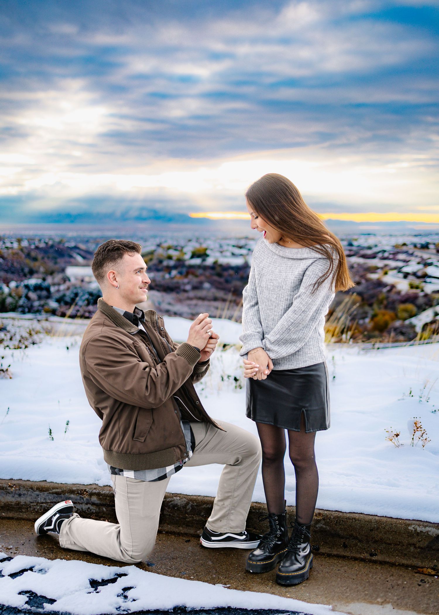 Man on one knee proposing to woman at snowy overlook with mountain city views at sunset.