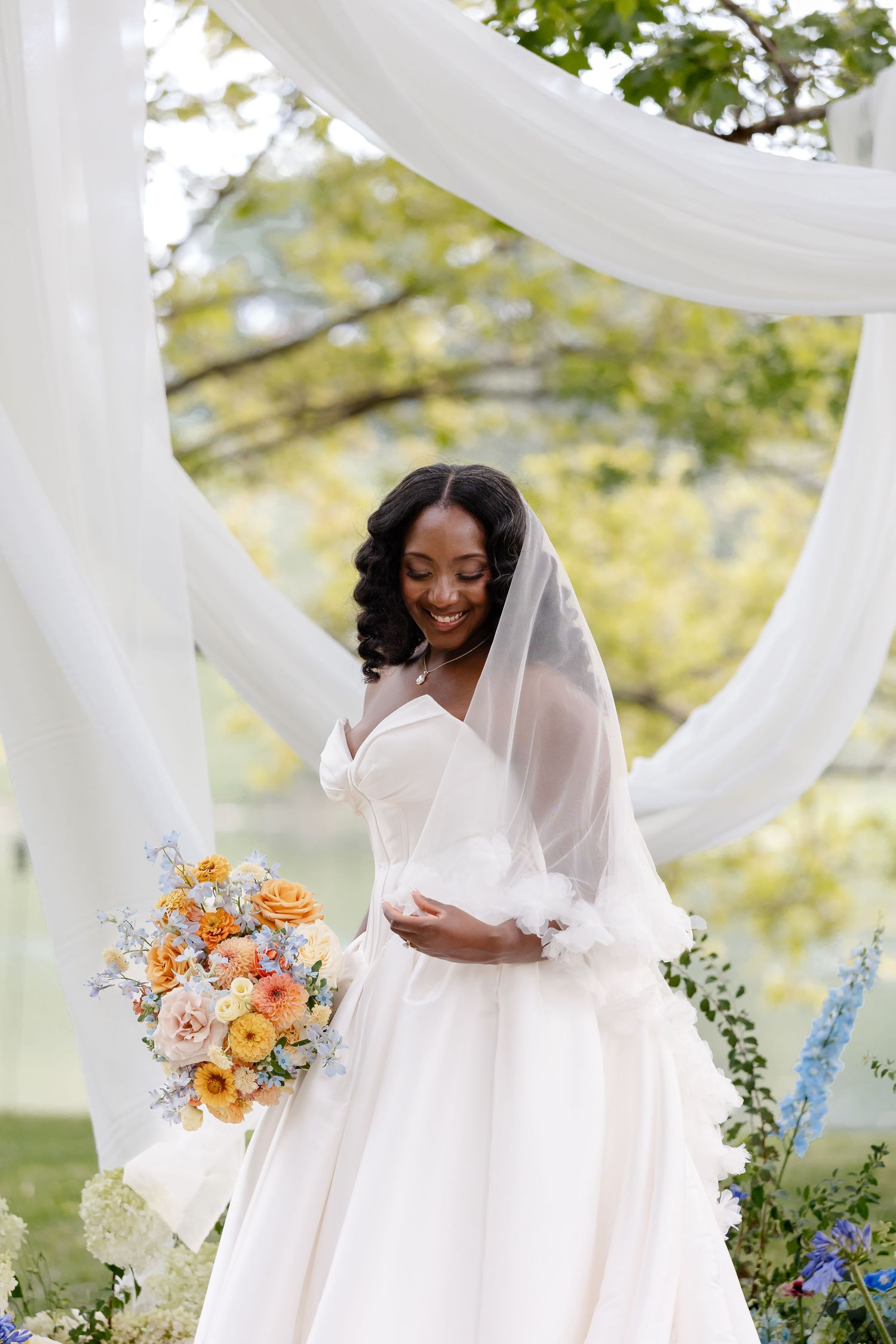Smiling bride in white gown holding orange bouquet under white draped arch outdoors.