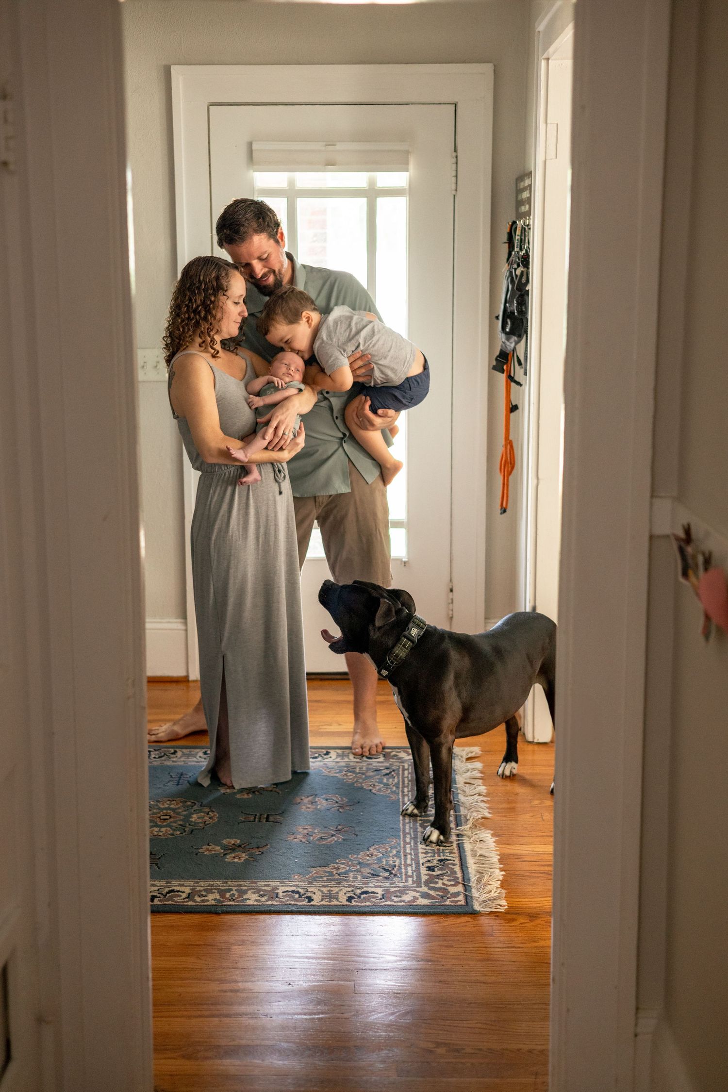 Family portrait with parents, two children, and black dog standing in a sunlit home hallway.