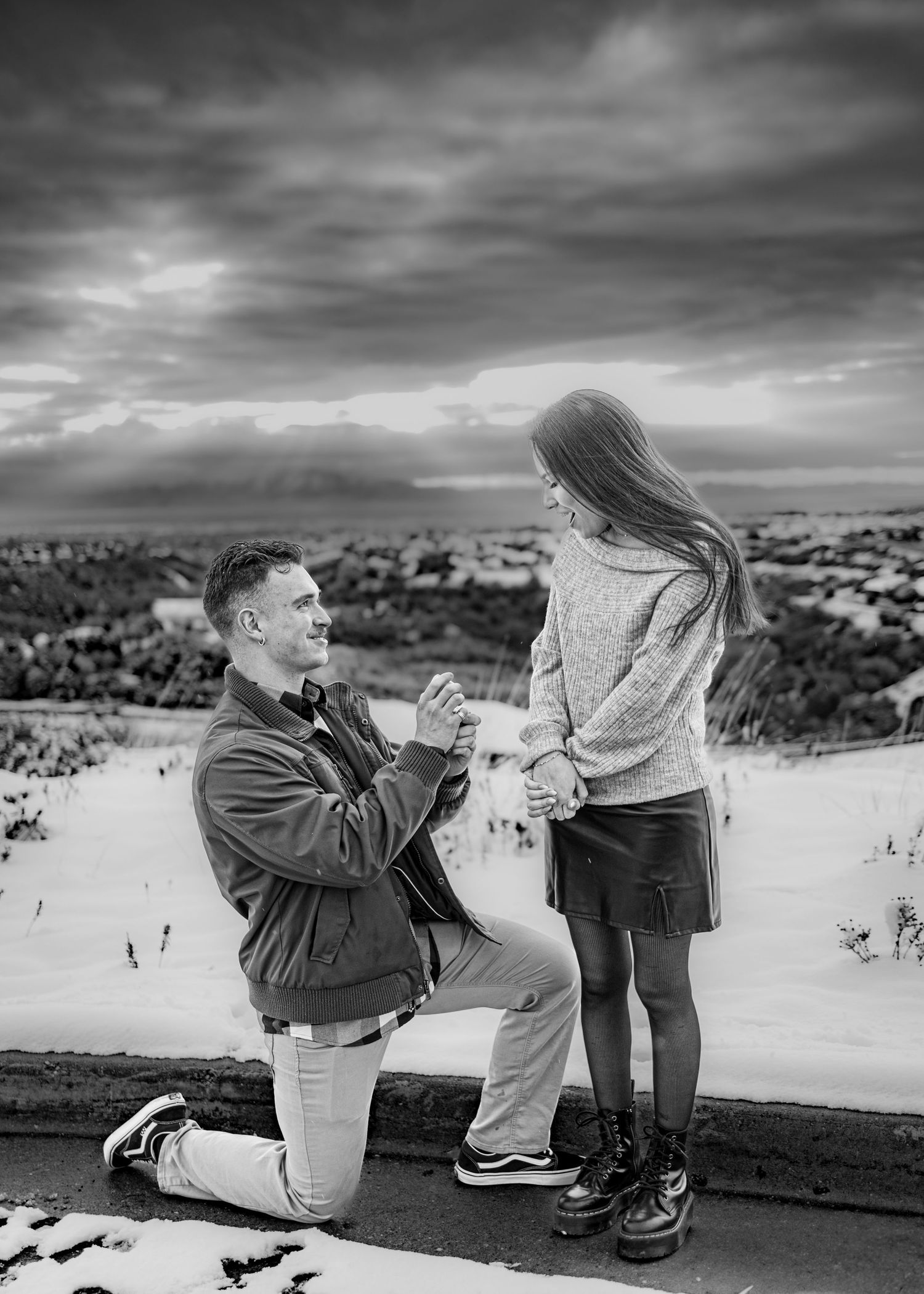 Black and white photo of a man on one knee proposing to a woman outdoors in a snowy winter landscape.