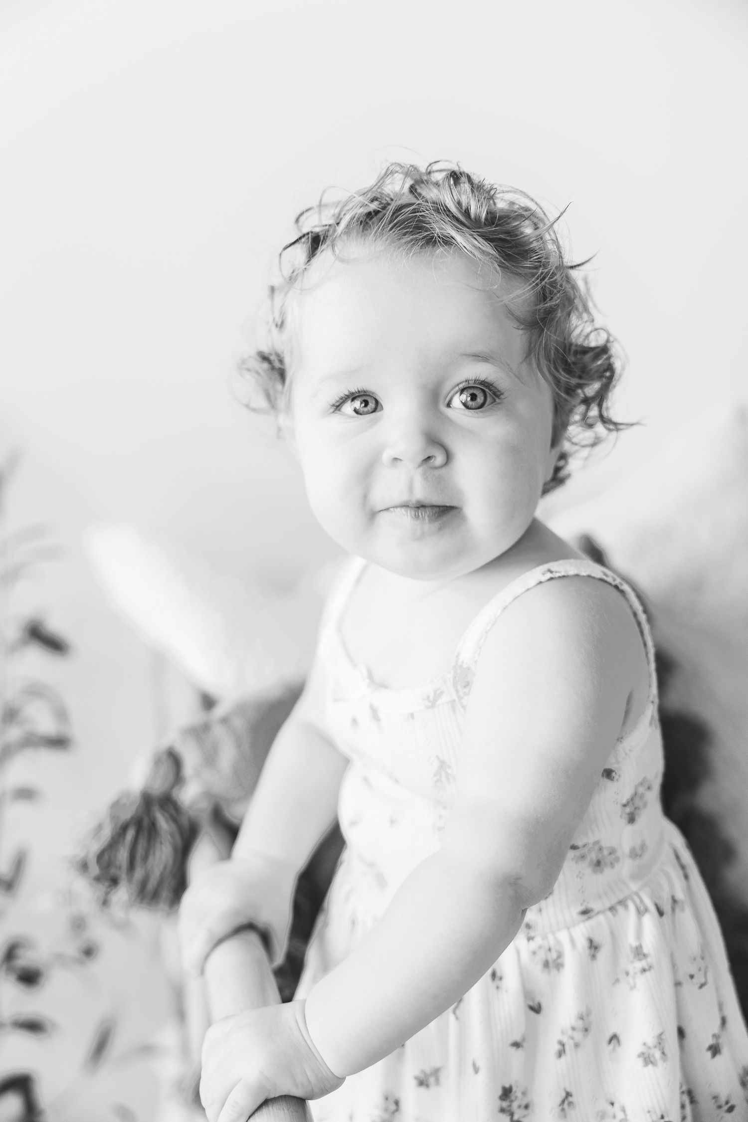Black and white portrait of a cute baby girl with curly hair wearing a floral dress, looking up with bright eyes.