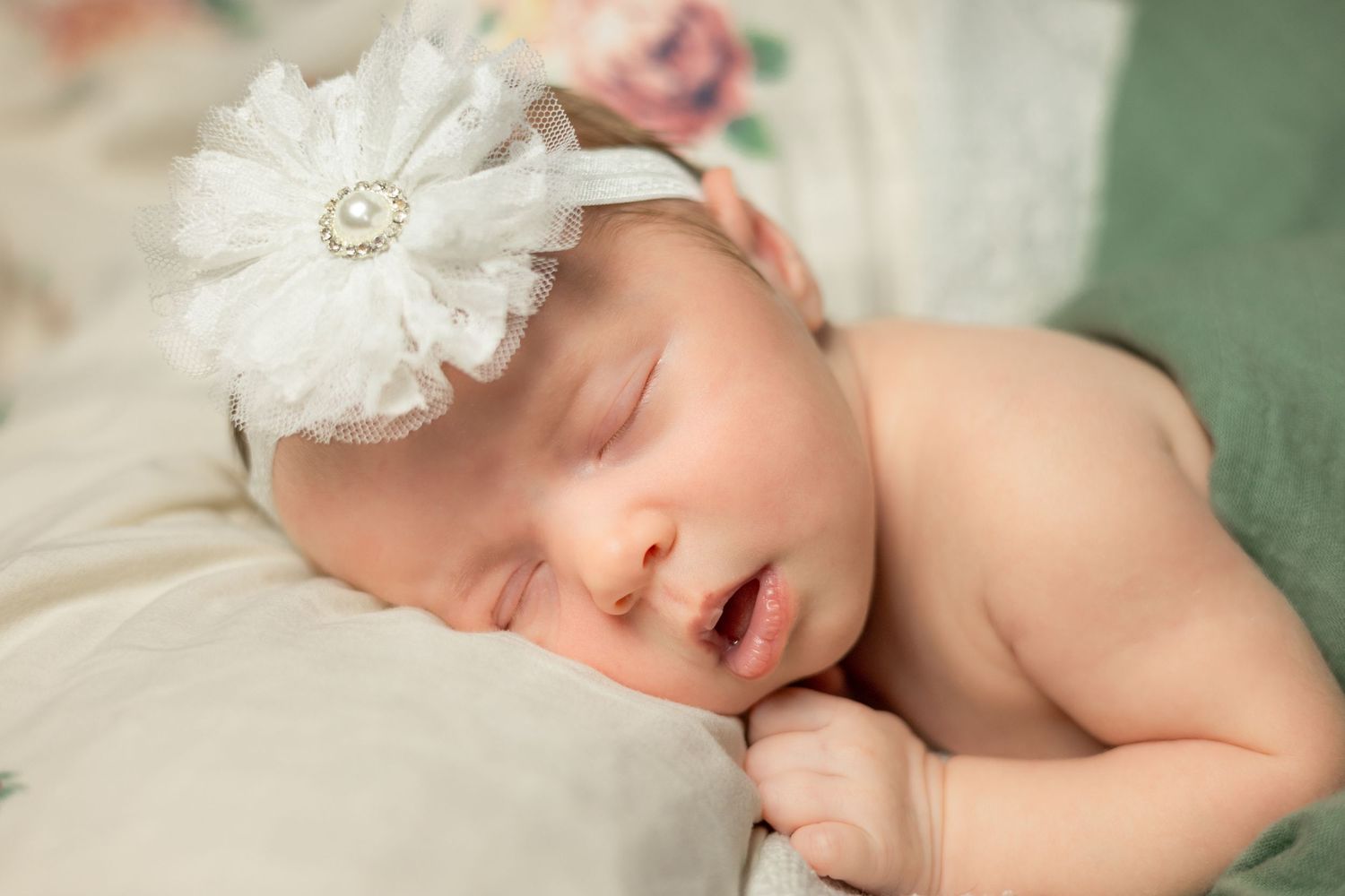 Adorable sleeping newborn baby girl wears white flower headband while resting on soft green blanket.