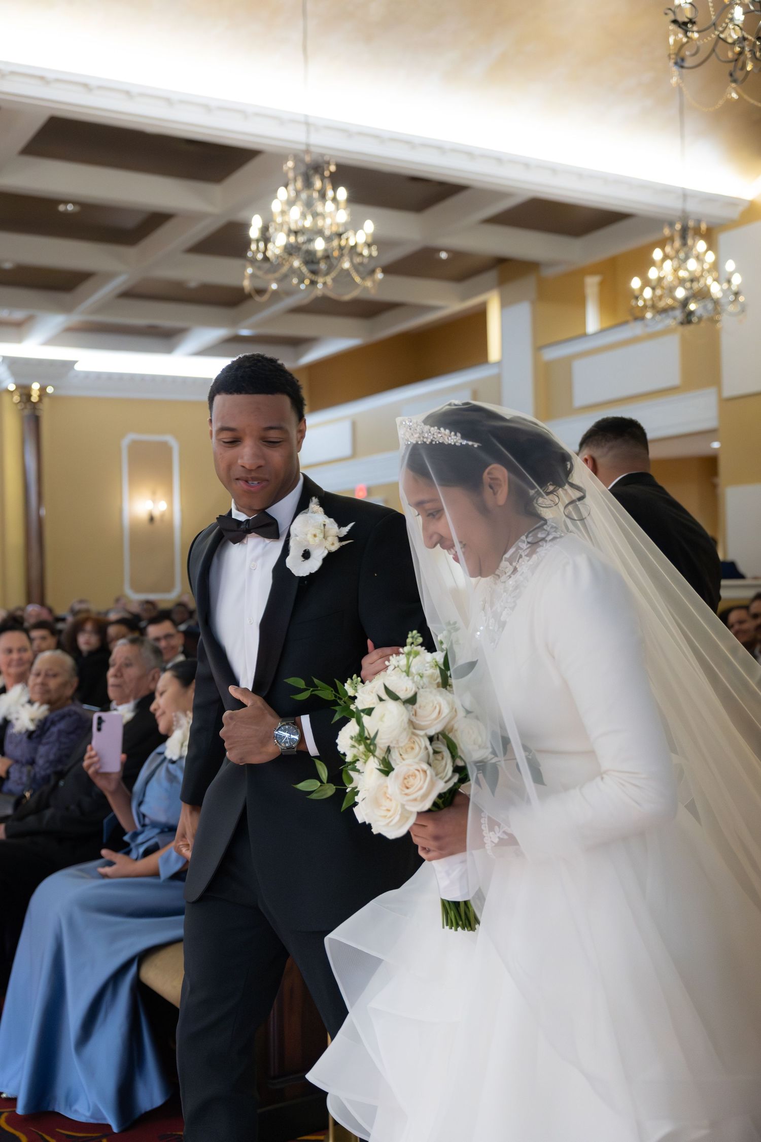 Interracial couple walking together at wedding ceremony, bride in lace gown holding white bouquet.