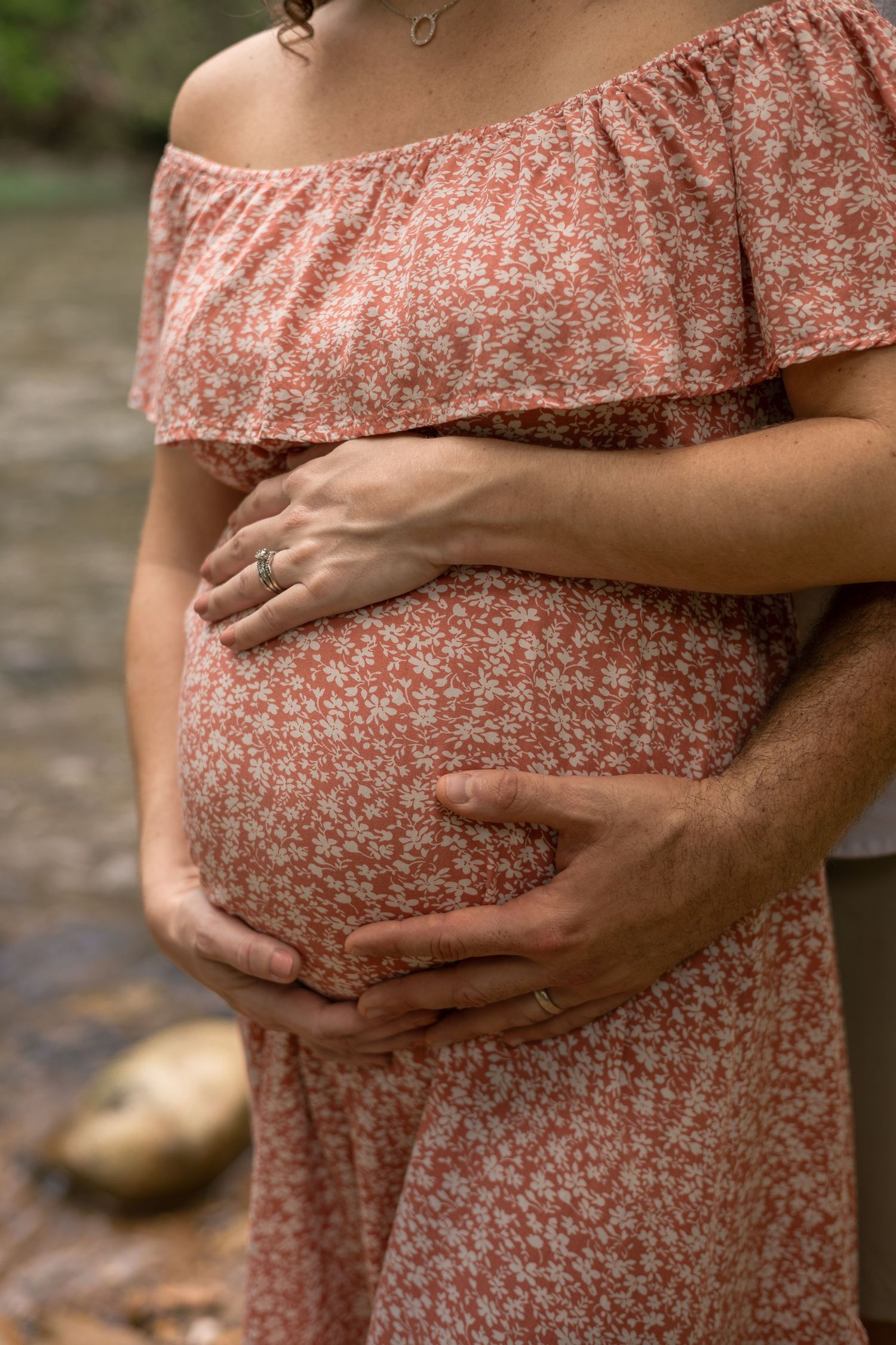 Pregnant woman in floral dress with partner's hands cradling baby bump near a creek.