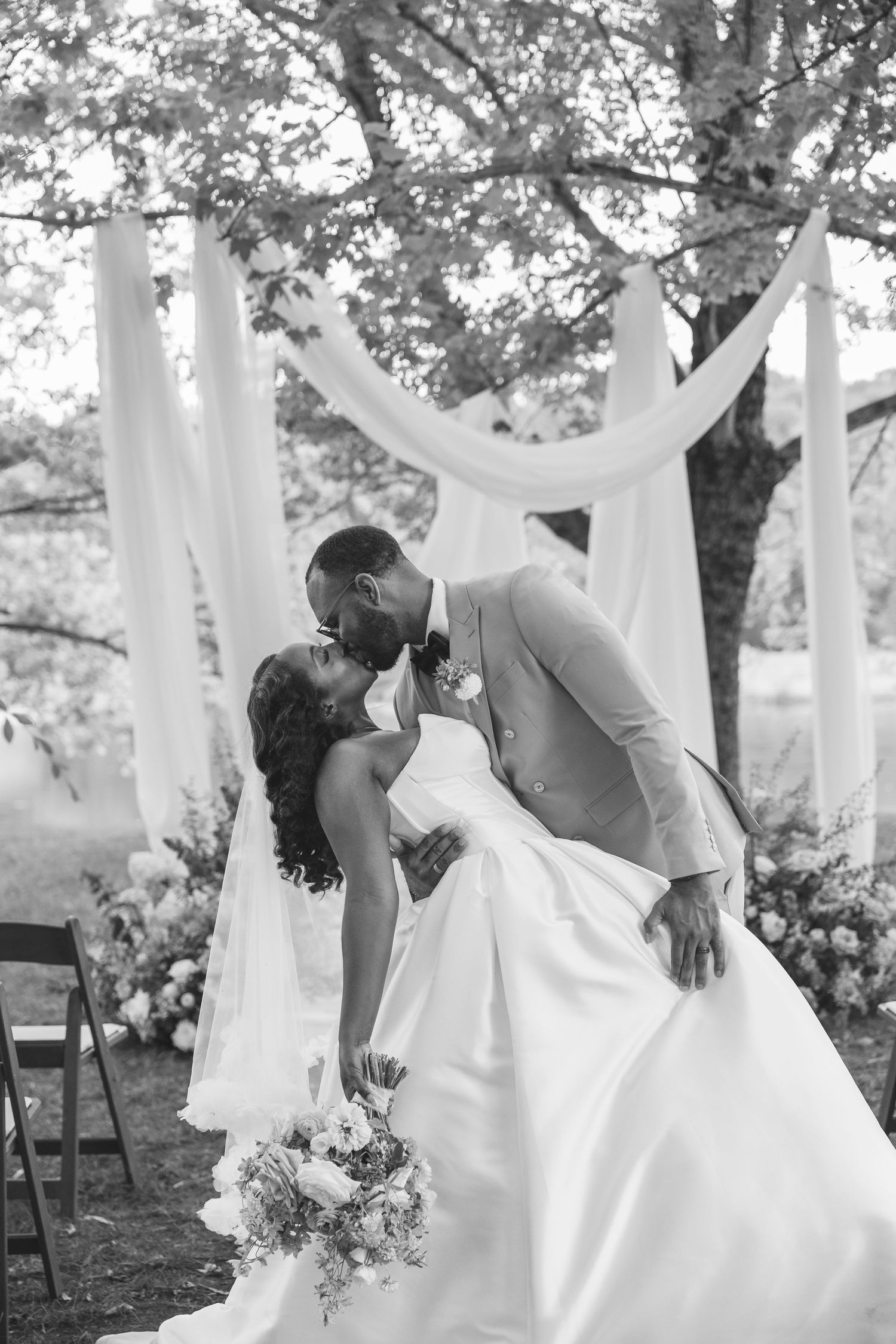 Black and white photo of a bride and groom sharing a kiss under a draped white fabric arch outdoors.