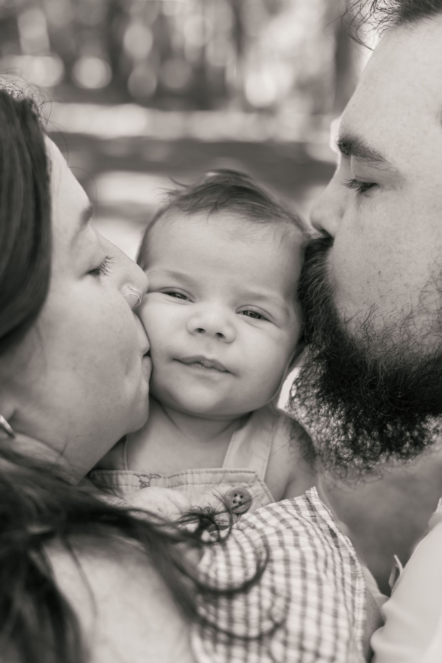 Black and white photo of parents kissing their baby on both cheeks outdoors.