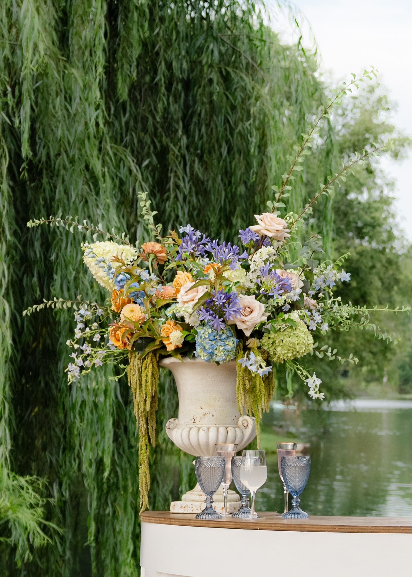 Elegant stone urn with lush floral arrangement of roses, hydrangeas, and wildflowers beside a serene willow tree pond.