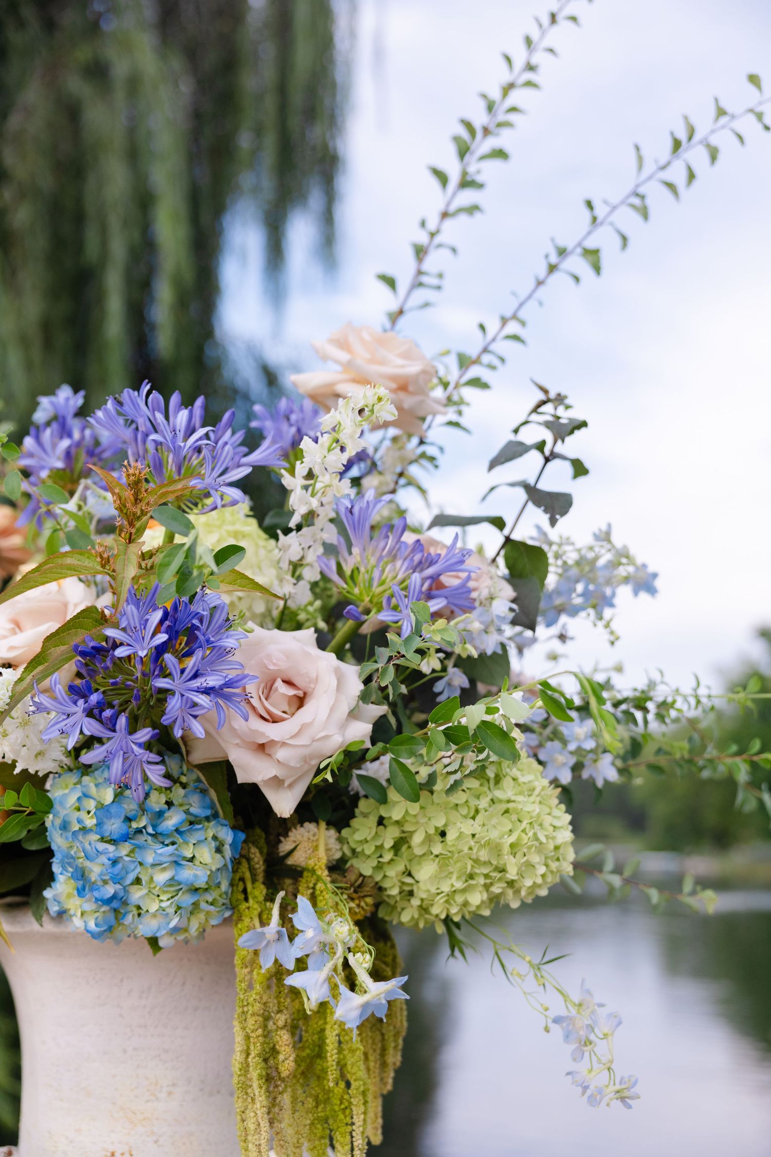 Elegant outdoor wedding floral arrangement featuring blue agapanthus, blush roses, and green hydrangeas in a white vase.