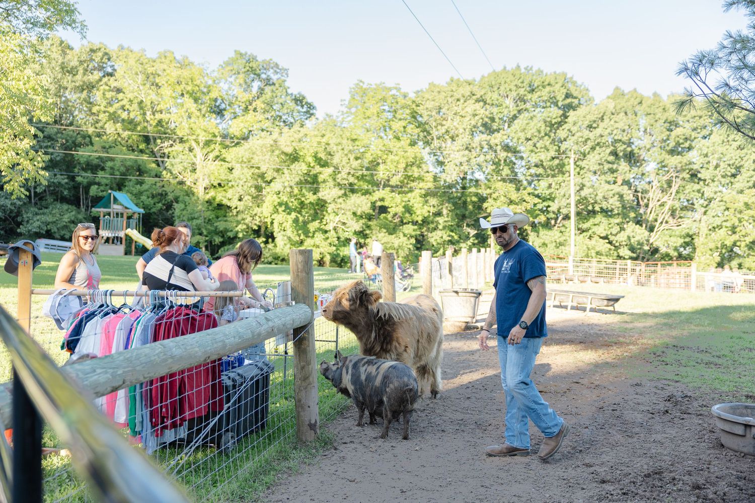 A farmer walks with sheep and a llama in a fenced farm area while visitors watch from behind a rail fence.