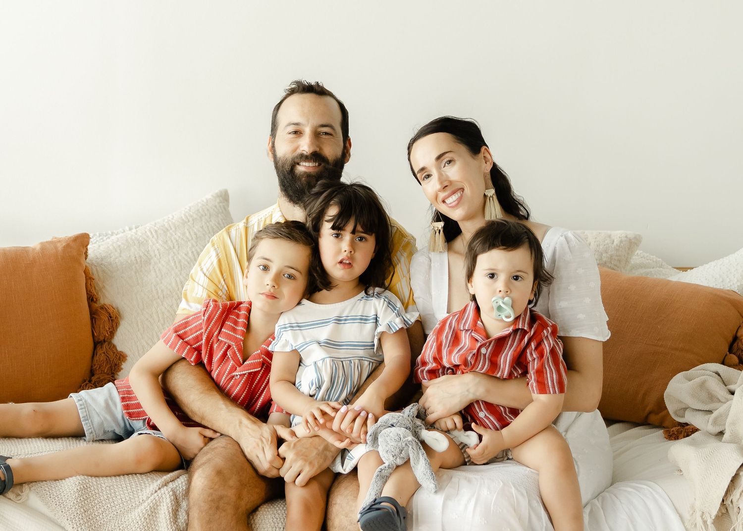 Bearded father, dark-haired mother, and three young children smiling together on cozy couch.
