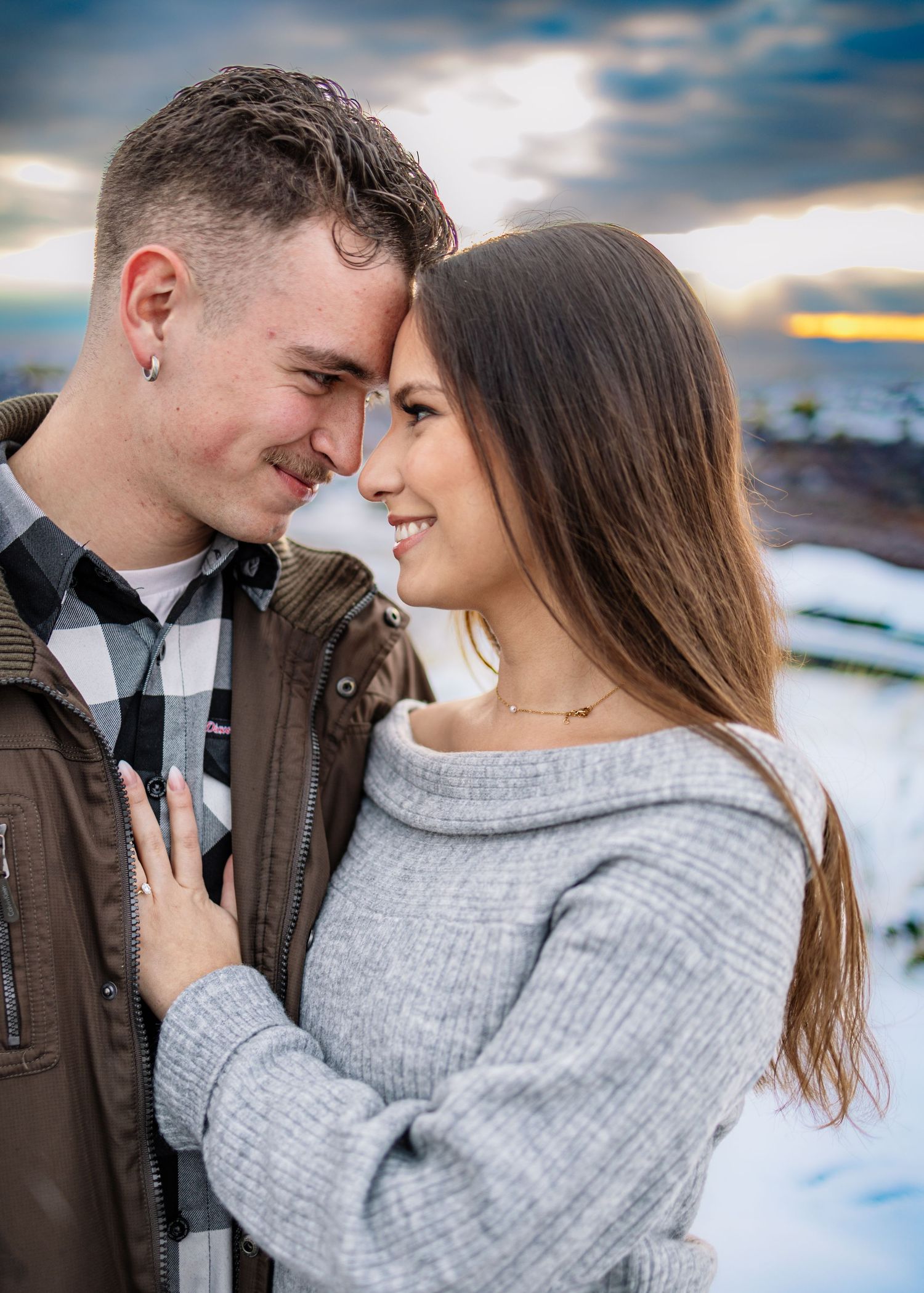 Happy engaged couple touching foreheads and smiling together against a stunning winter sunset sky.
