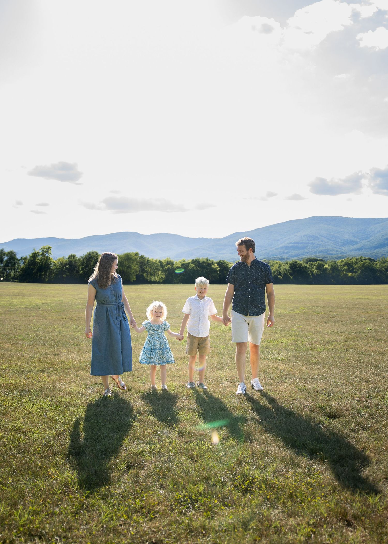 Family of four holds hands in a sunny green field with mountain views in the background.
