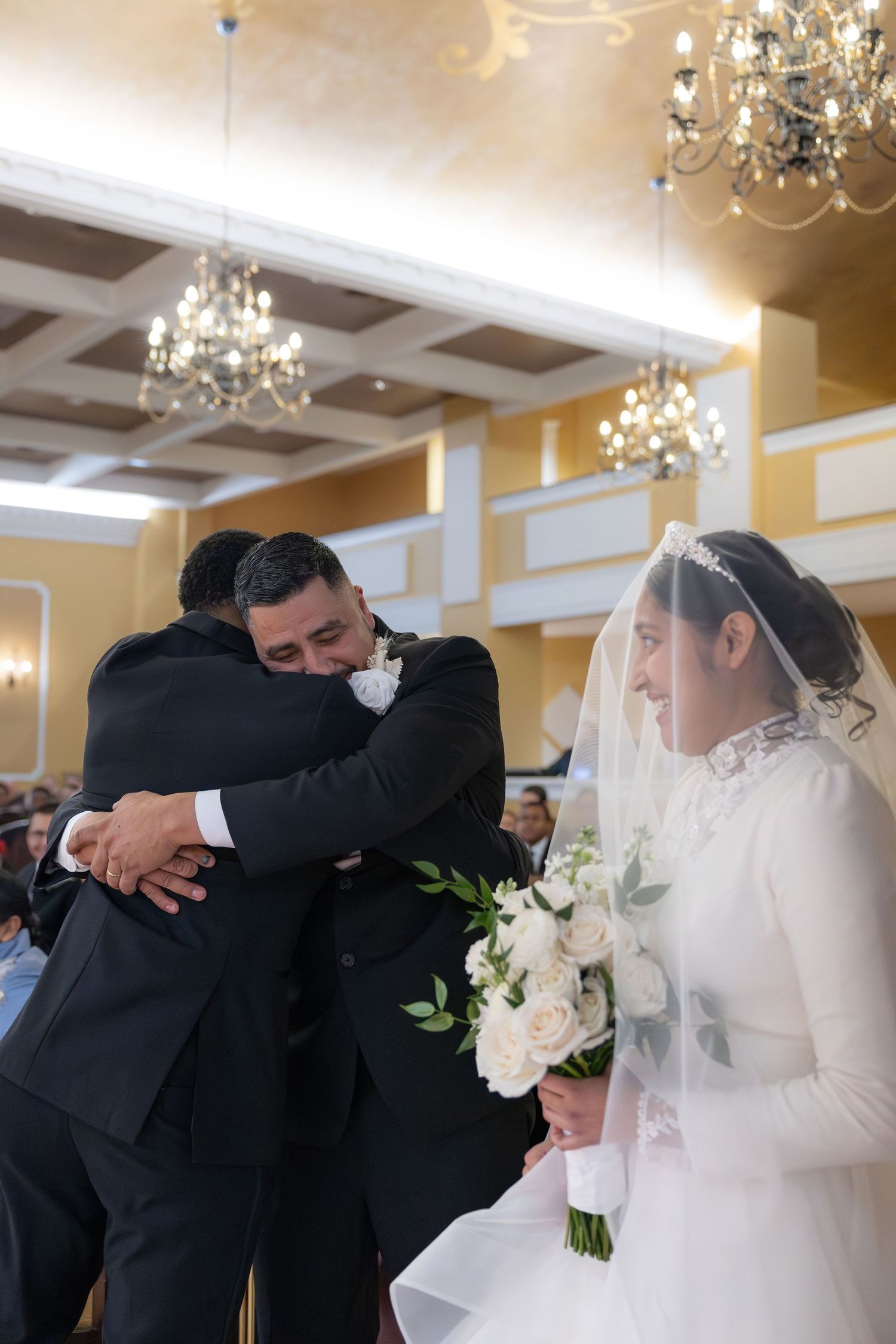 Groom embracing father-in-law as bride in white veil holds bouquet during wedding ceremony.