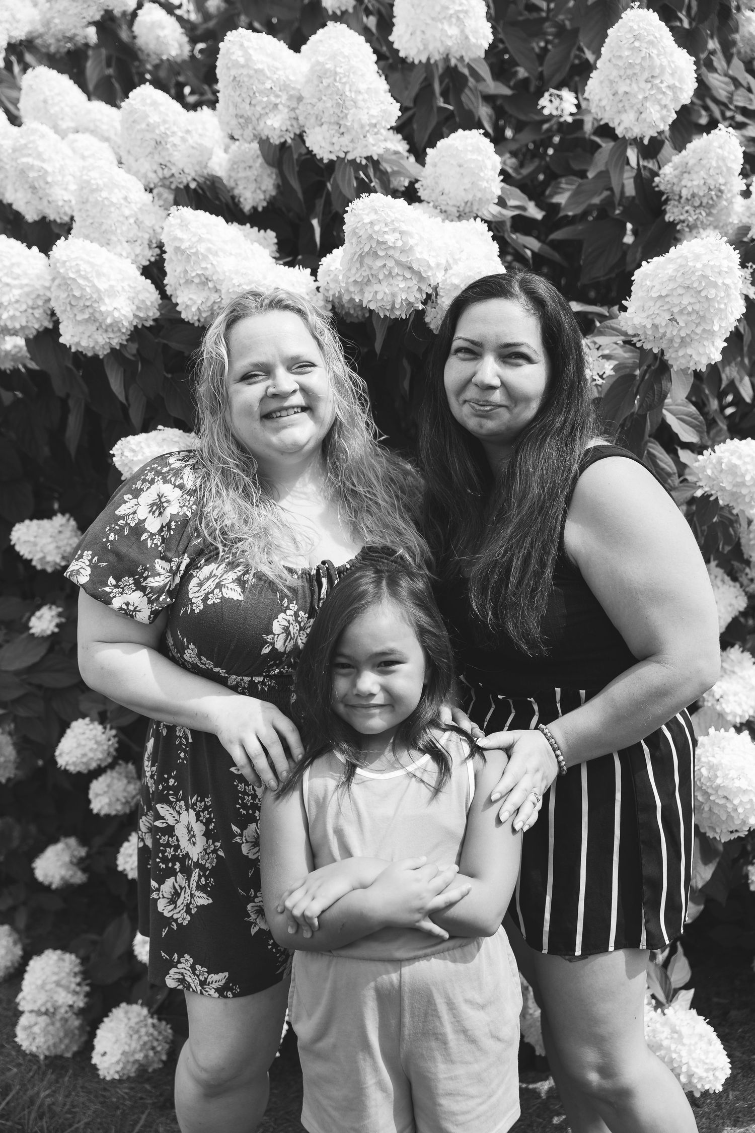 Black and white photo of two women and a young girl posing in front of blooming white hydrangea bushes outdoors.