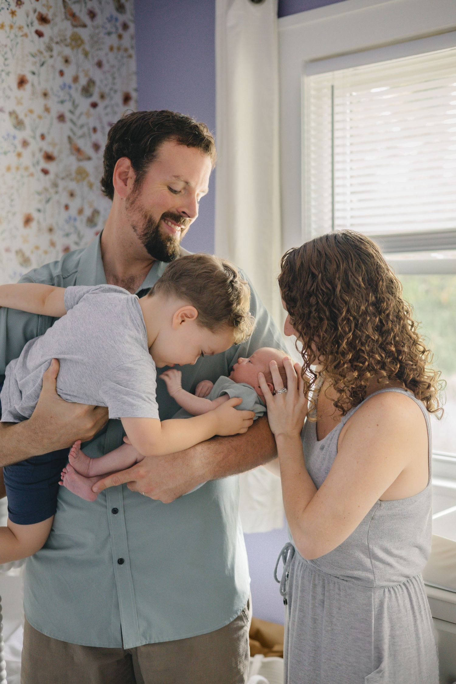 Smiling family with toddler boy and newborn baby shares tender moment together in decorated nursery.