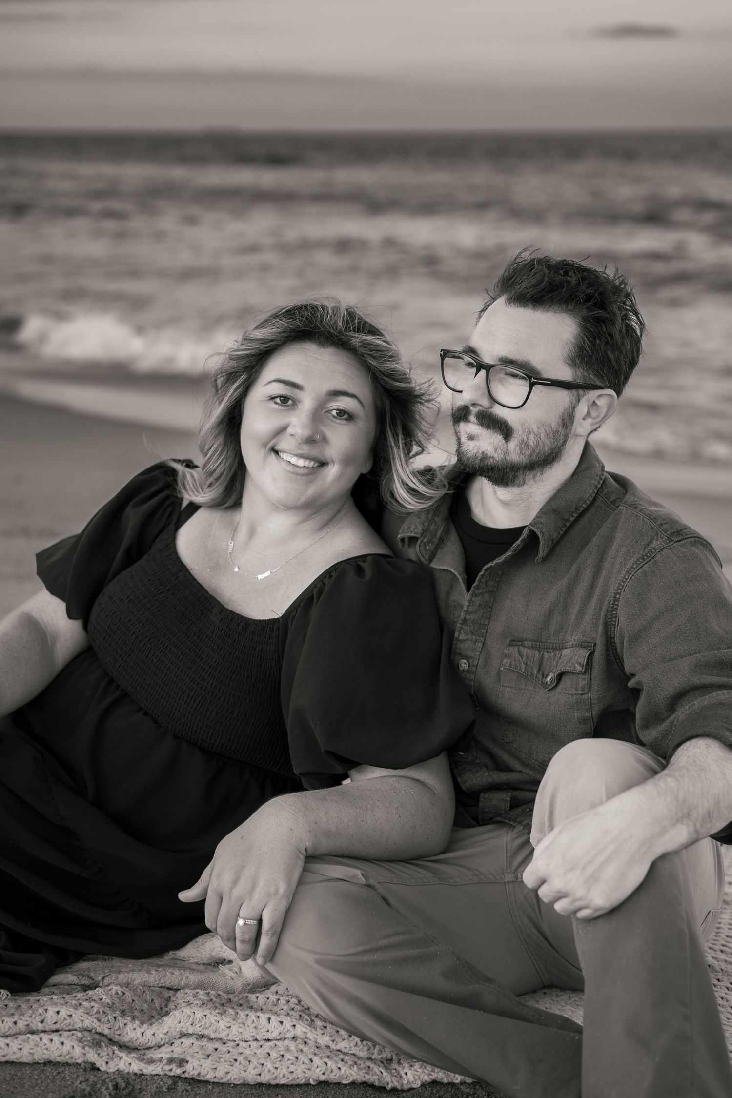Black and white photo of a smiling couple sitting together on a beach, with ocean waves in the background.