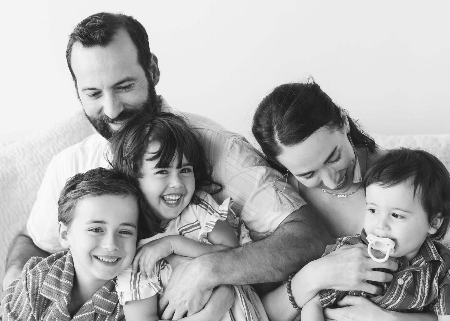 Black and white family portrait of parents with three young children smiling and cuddling together on a couch.