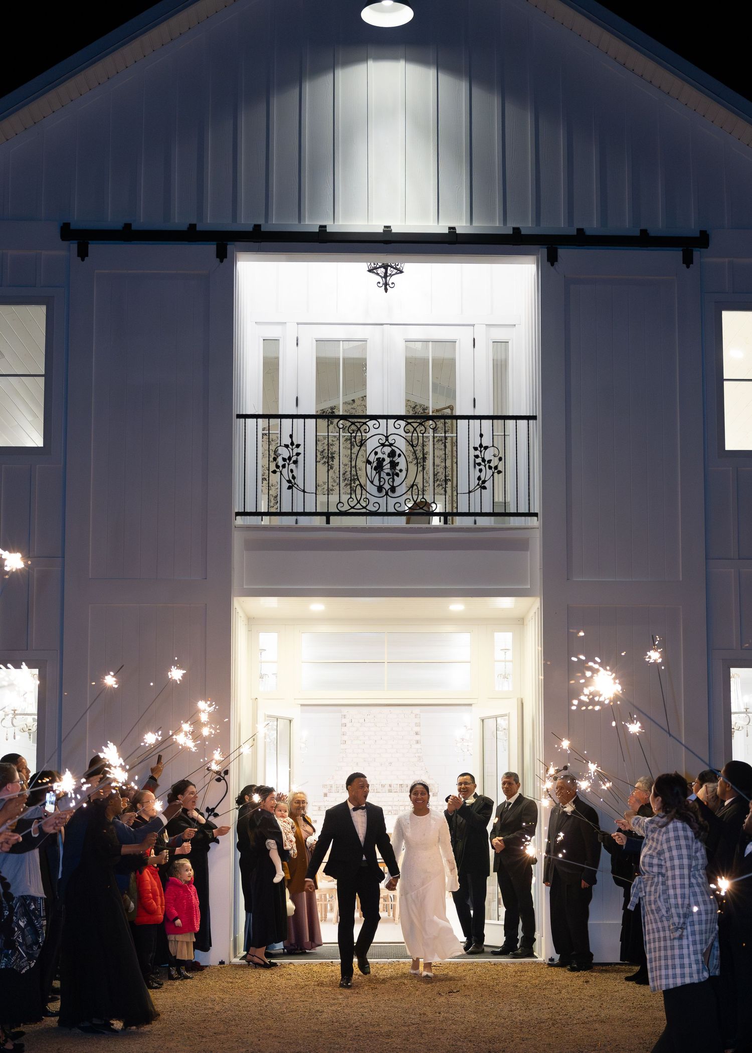 Bride and groom exit white barn wedding venue through sparkler send-off tunnel.