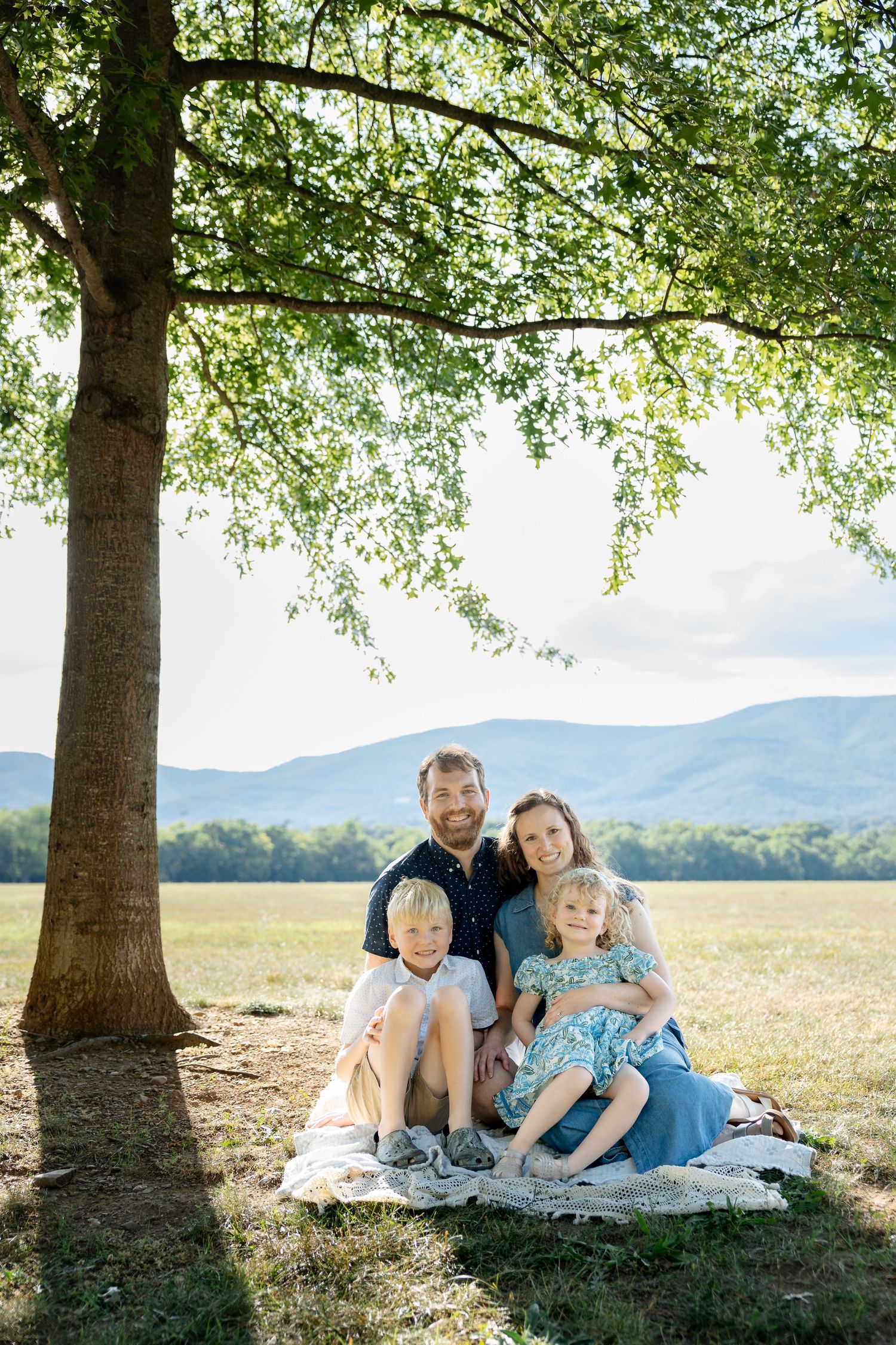 Family of four sitting on a blanket under a large tree with mountain views behind them.