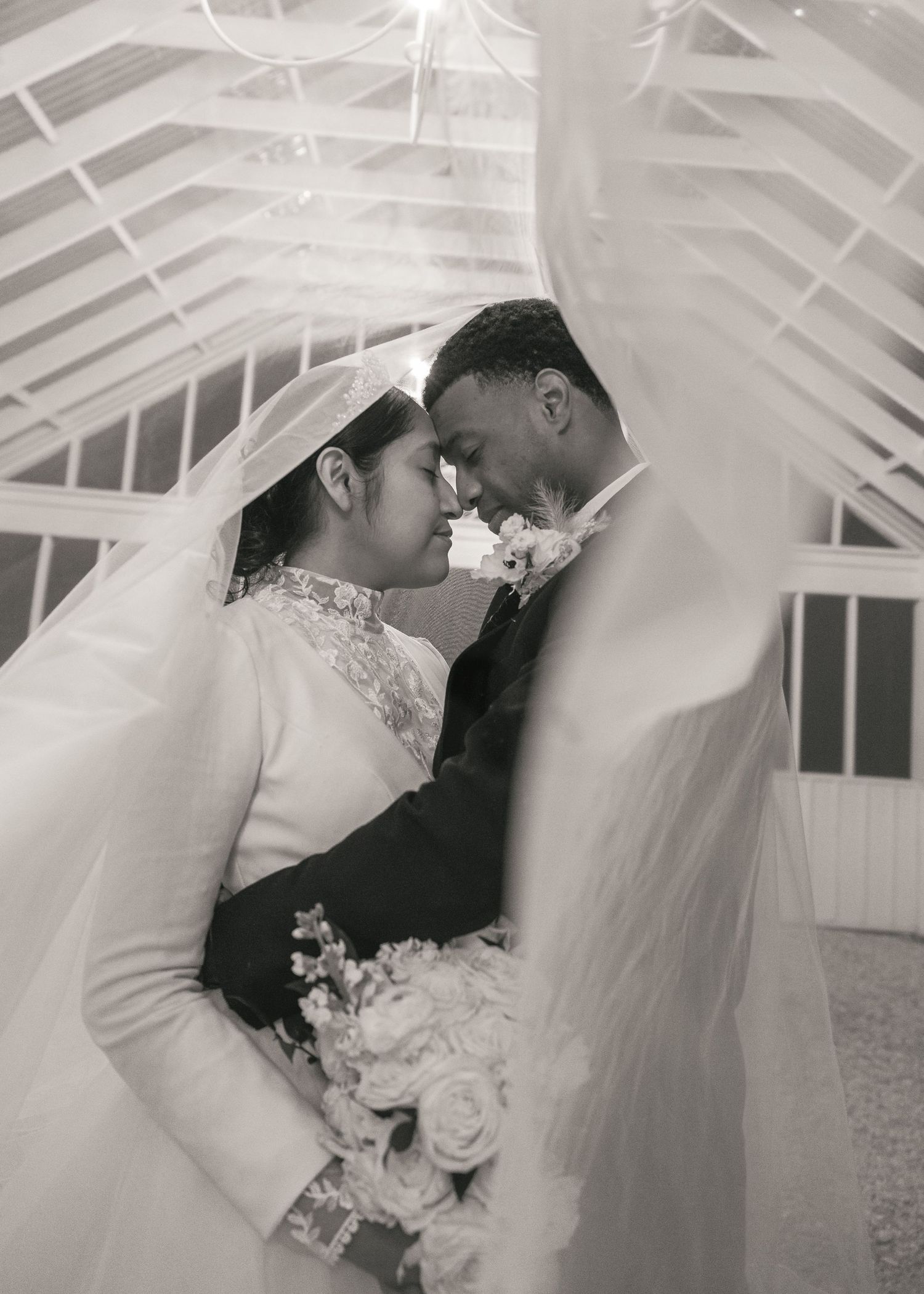 Black and white wedding photo of bride and groom touching foreheads tenderly, framed by flowing veil in elegant venue.
