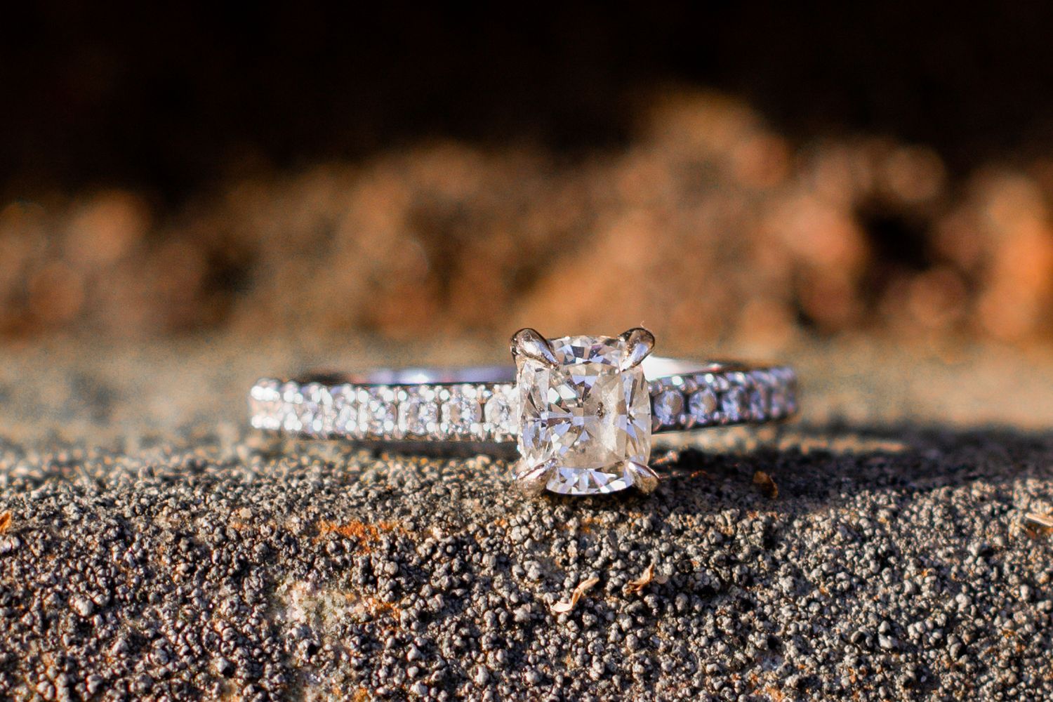 Close-up of a stunning cushion-cut diamond engagement ring with pavé band on a rustic surface.