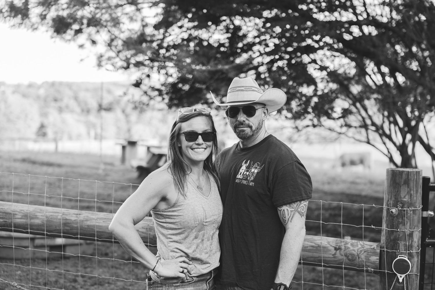 Black and white photo of a smiling couple posing by a farm fence, woman in sunglasses and man in cowboy hat.