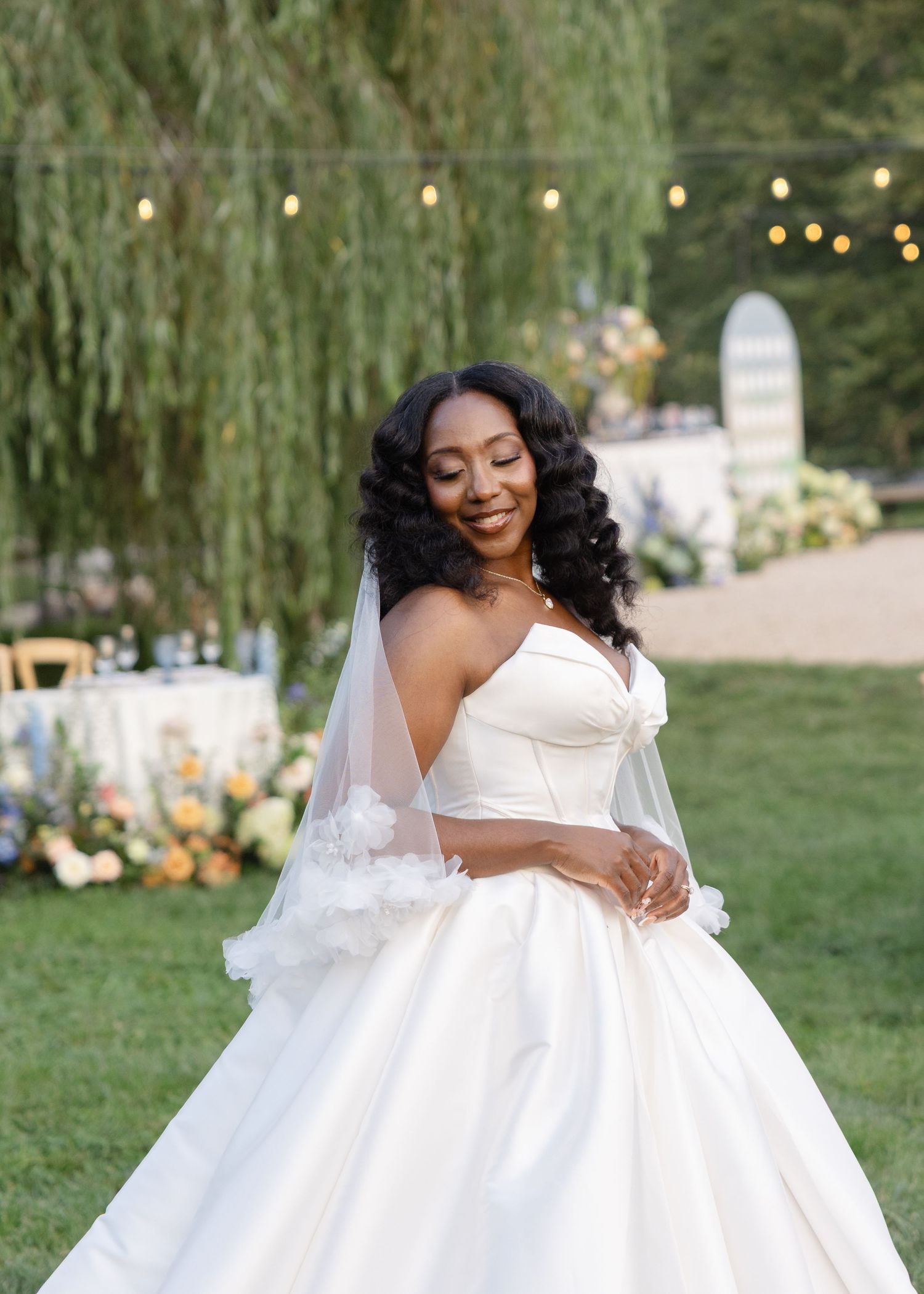 Radiant Black bride in a strapless ball gown with floral veil twirling outdoors under willow trees with string lights.