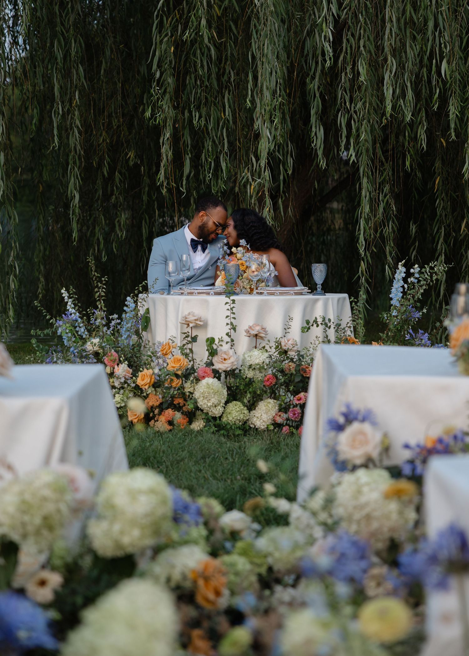 Bride and groom kissing at sweetheart table surrounded by lush colorful floral arrangements under a weeping willow tree.