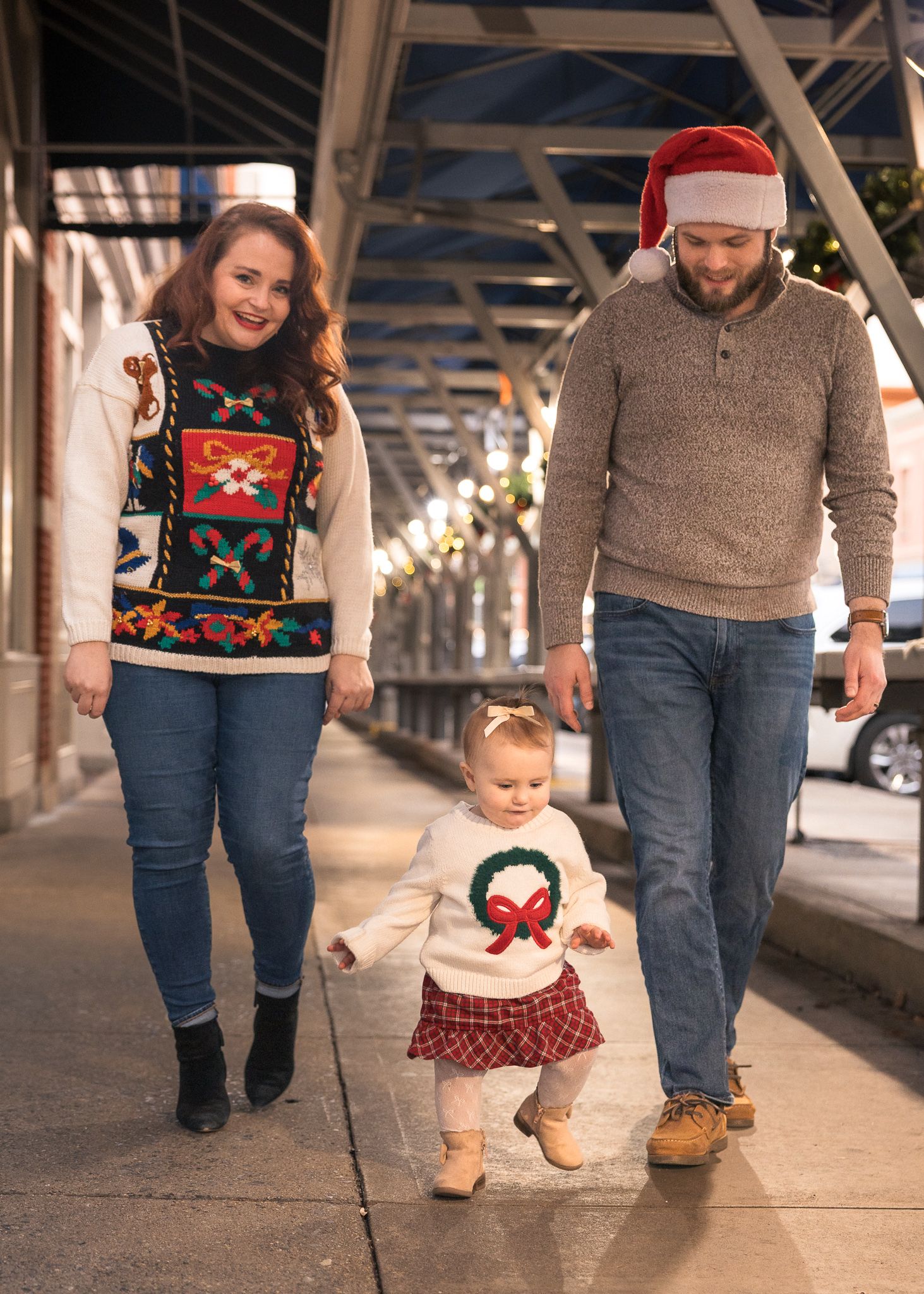 Family in Christmas sweaters walking on a sidewalk with toddler in holiday outfit and dad wearing Santa hat.