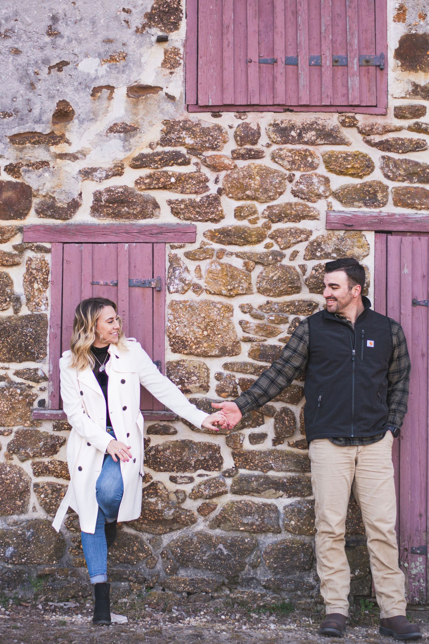 Couple holding hands and smiling at each other against a rustic stone wall with purple shutters.