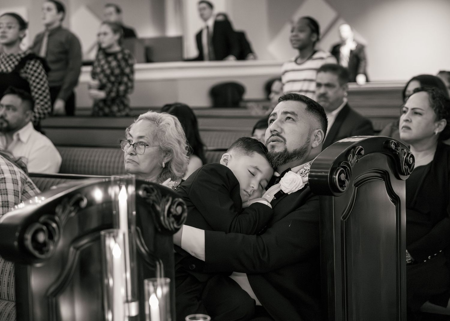 Black and white photo of a child sleeping on a mans lap during a church service in wooden pews.