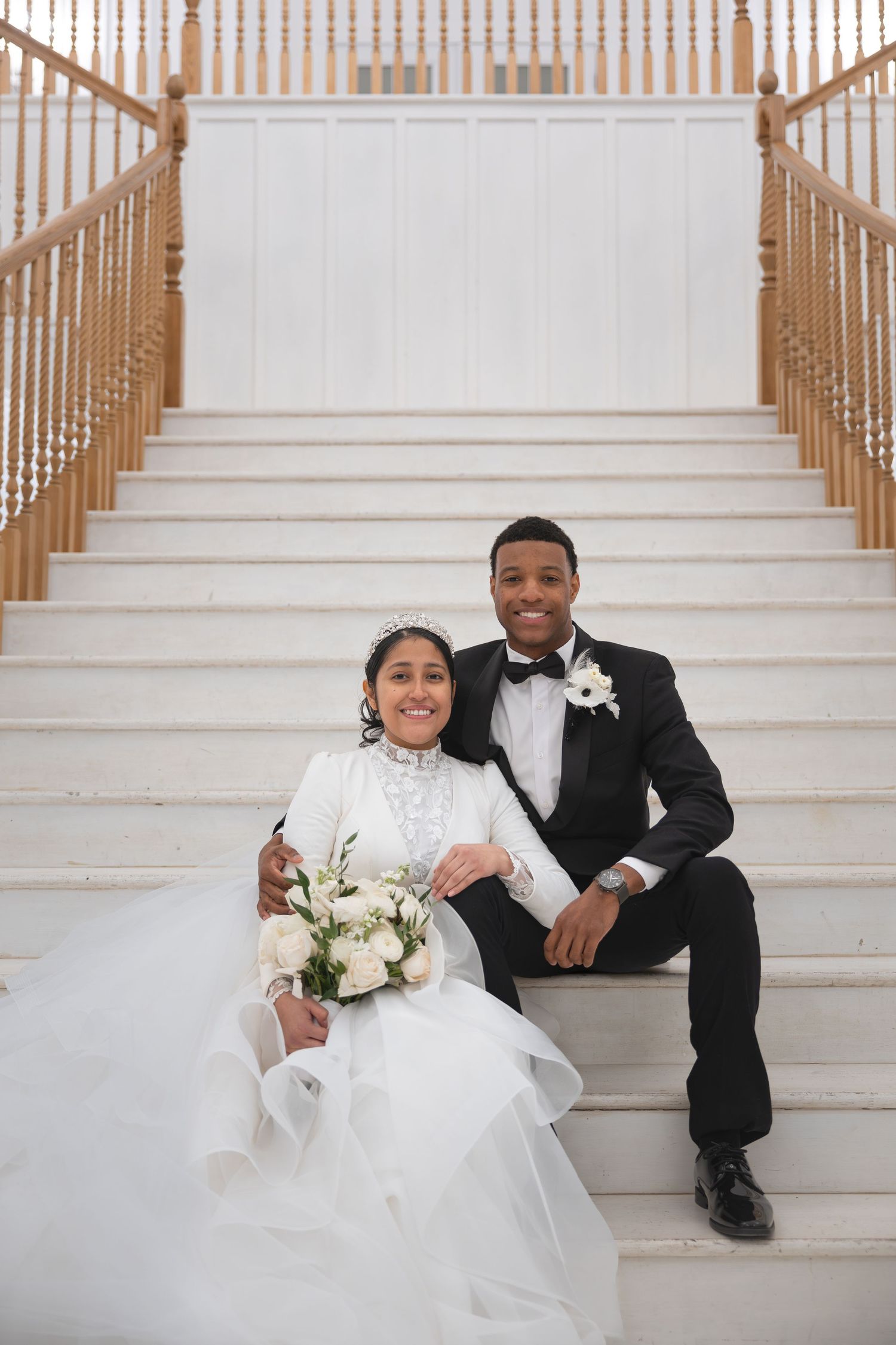 Newlywed couple sitting on grand staircase, bride in white gown holding bouquet, groom in tuxedo.