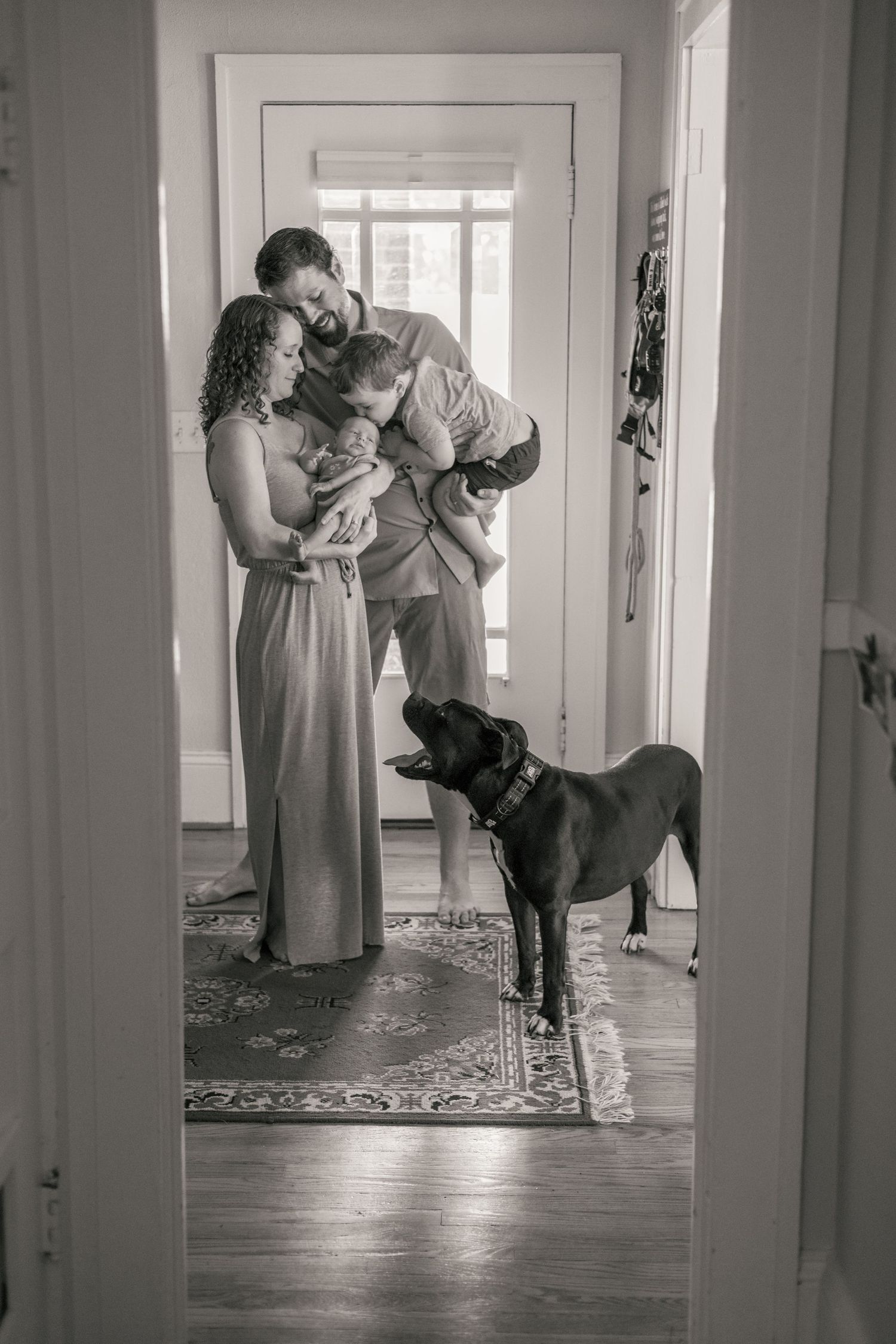 Black and white family portrait with parents holding two children and a Labrador dog standing in a sunlit hallway.