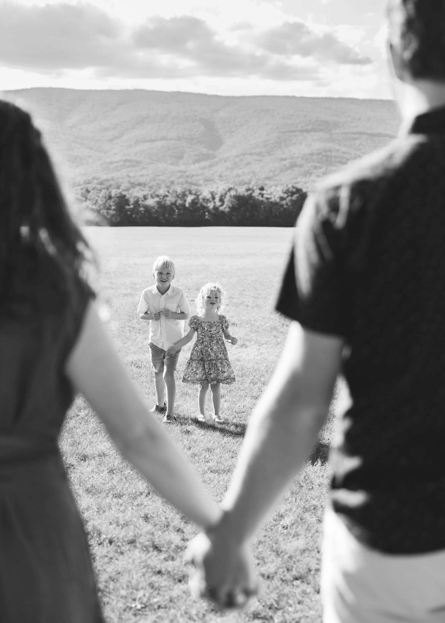 Black and white photo of parents holding hands watching two young children run across an open field with mountains behind.
