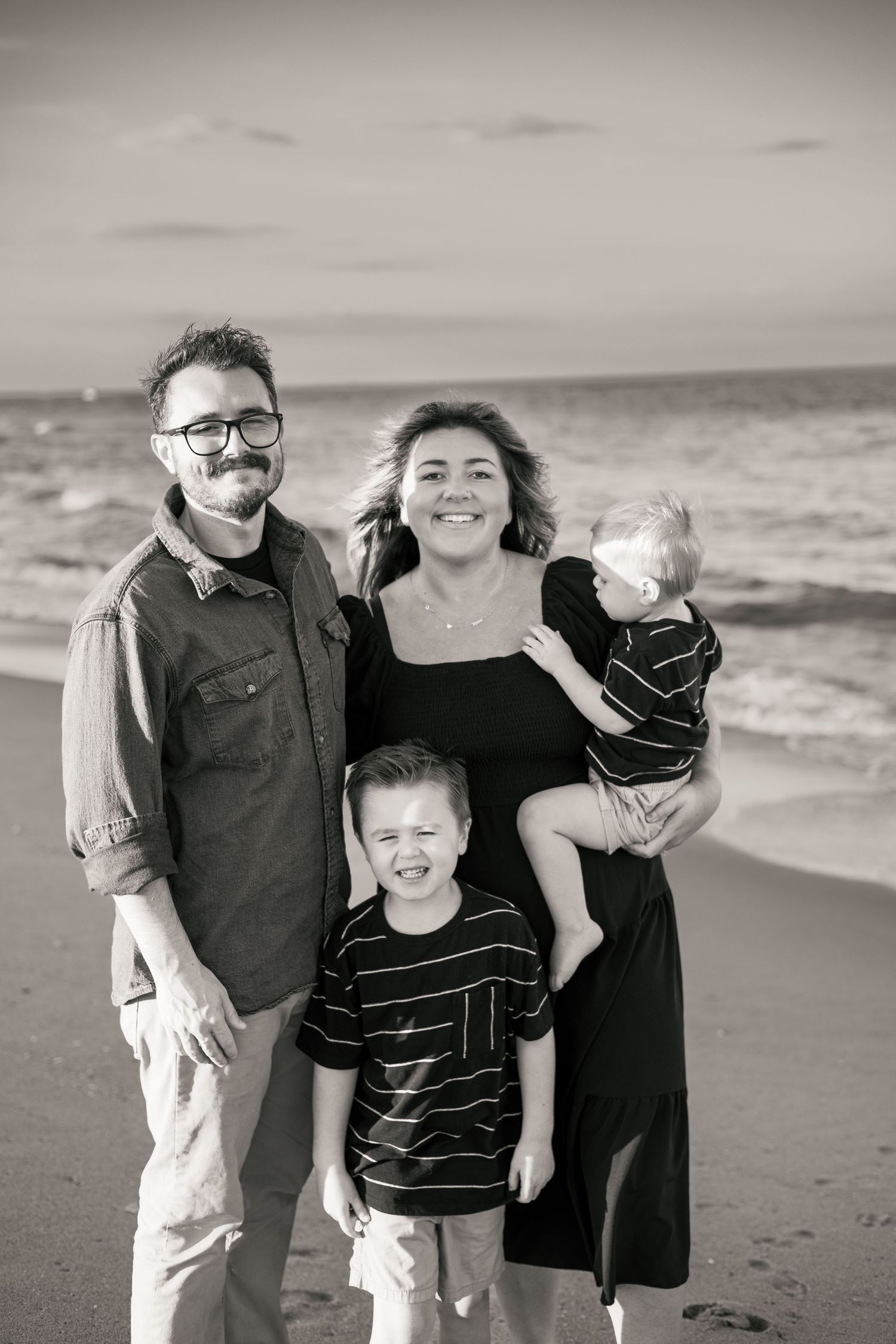 Black and white family portrait of parents with two young boys smiling on a beach with ocean waves behind them.