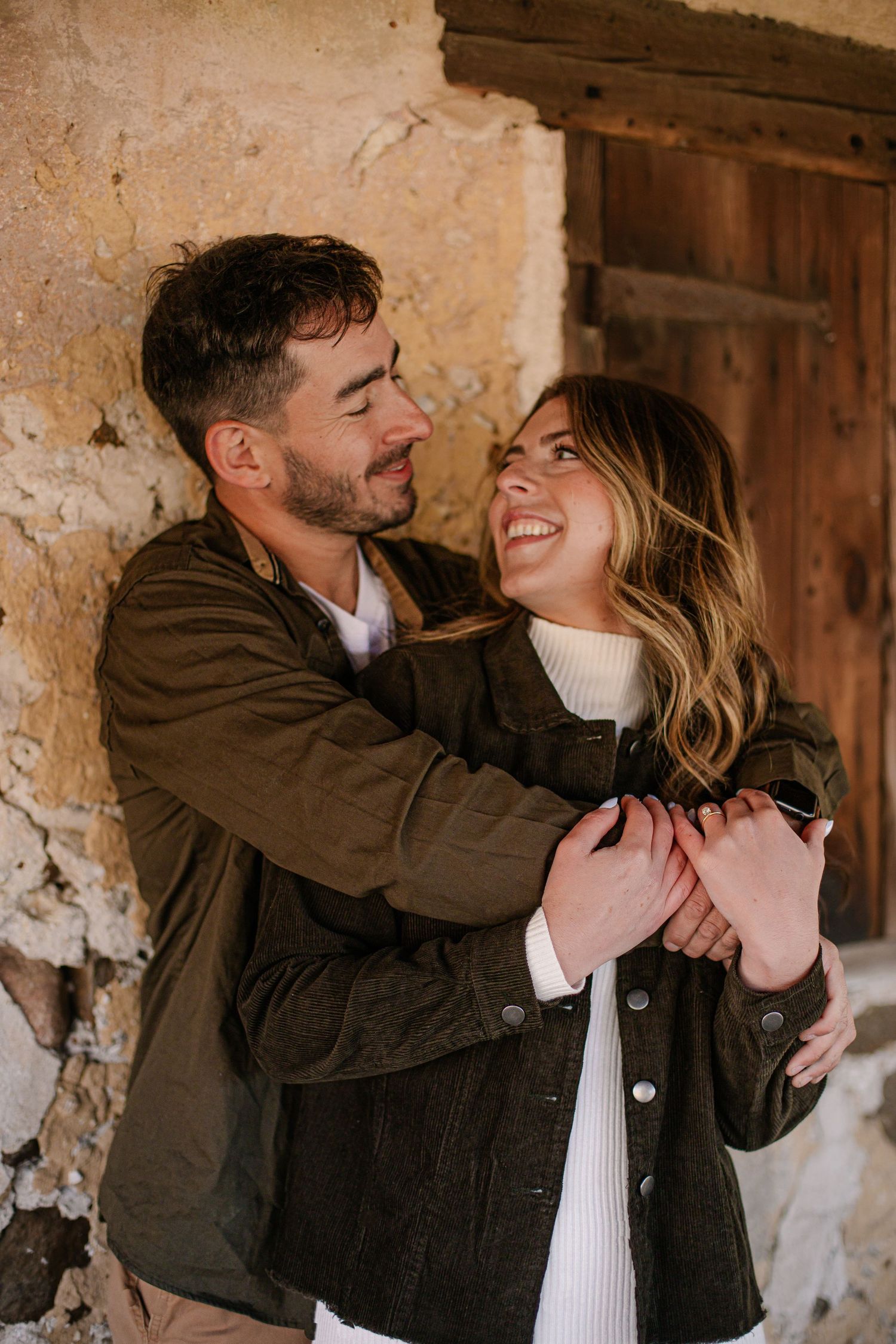 Couple sharing a tender embrace and smile against a rustic stone wall with wooden door.
