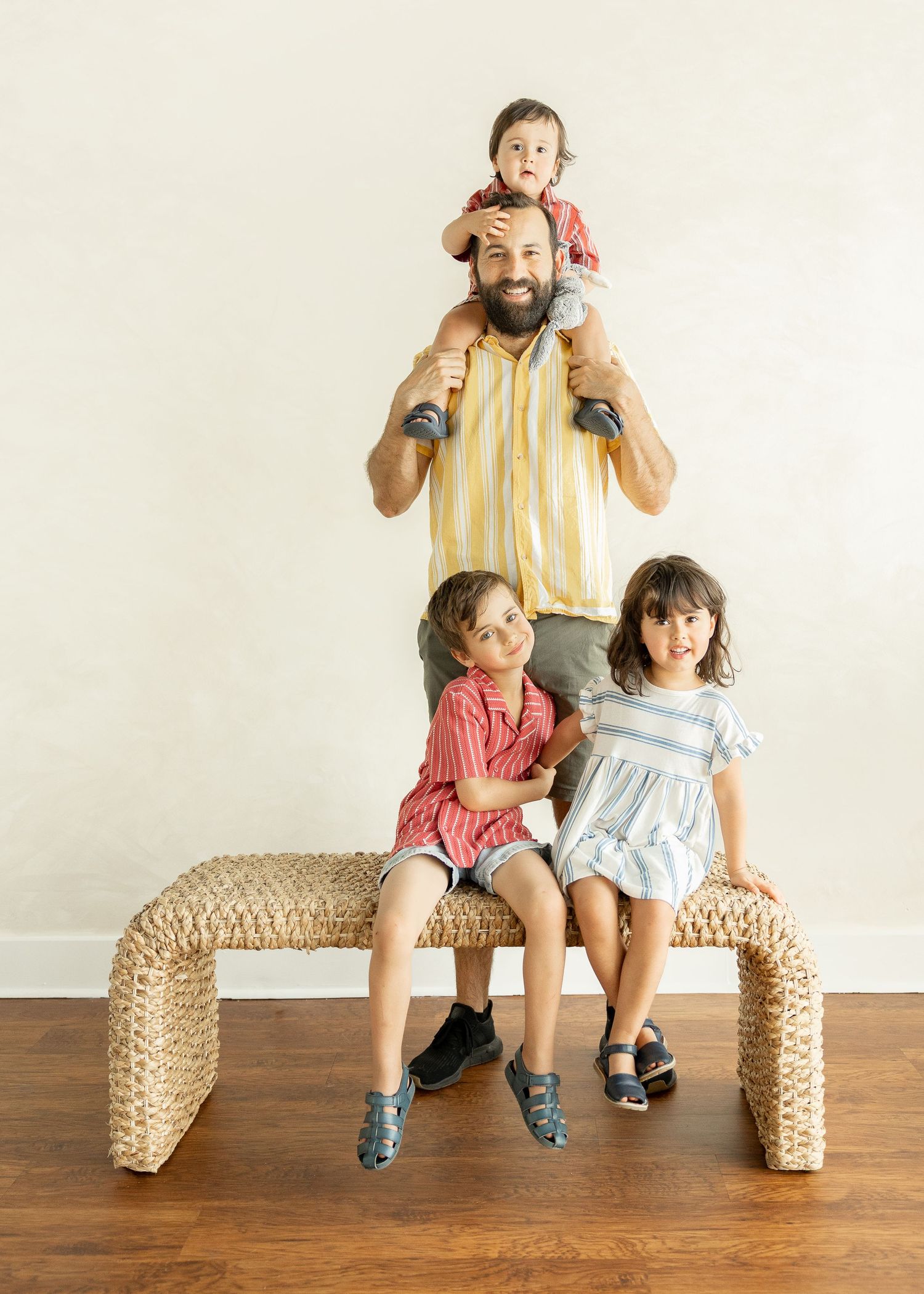 Bearded father with toddler on shoulders and two kids sitting on woven bench indoors.