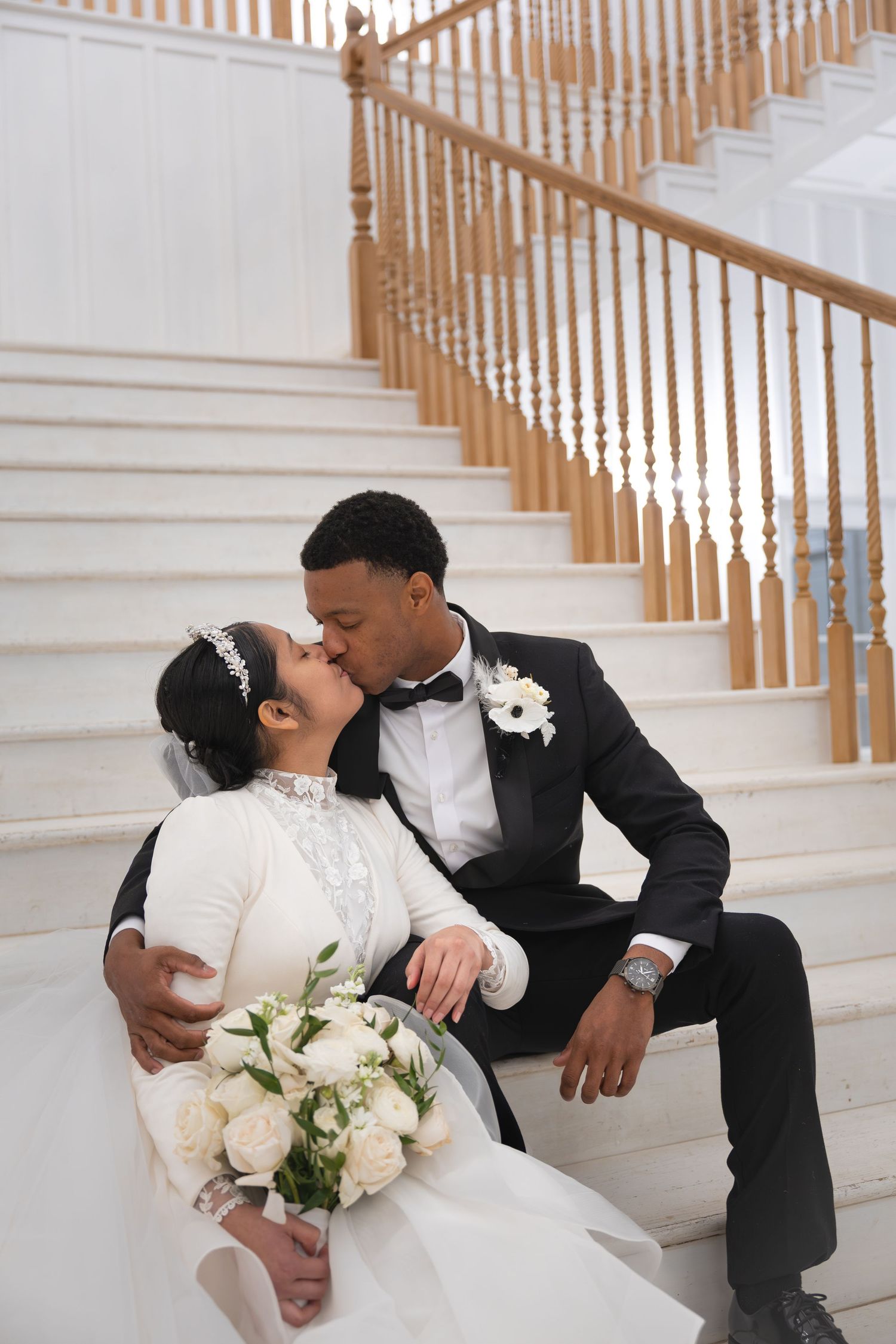 Bride and groom share romantic kiss on grand staircase with white bouquet.