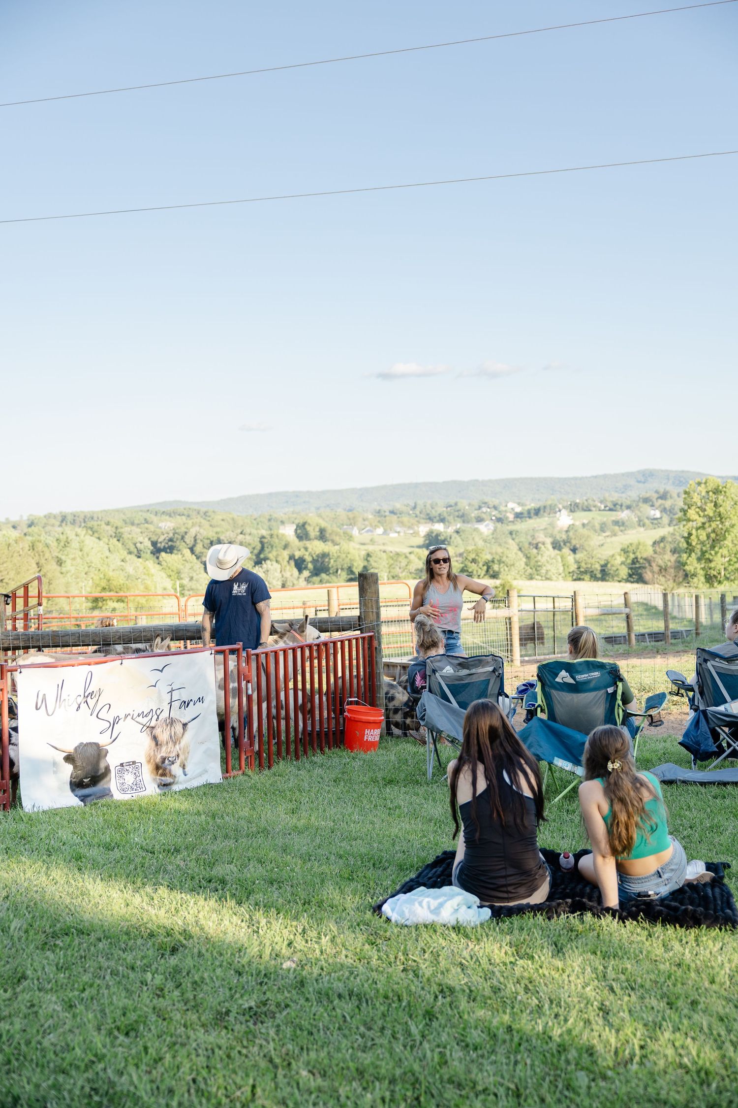 People relaxing on grass at a rural farm event with rolling hills and a decorative longhorn cattle sign.
