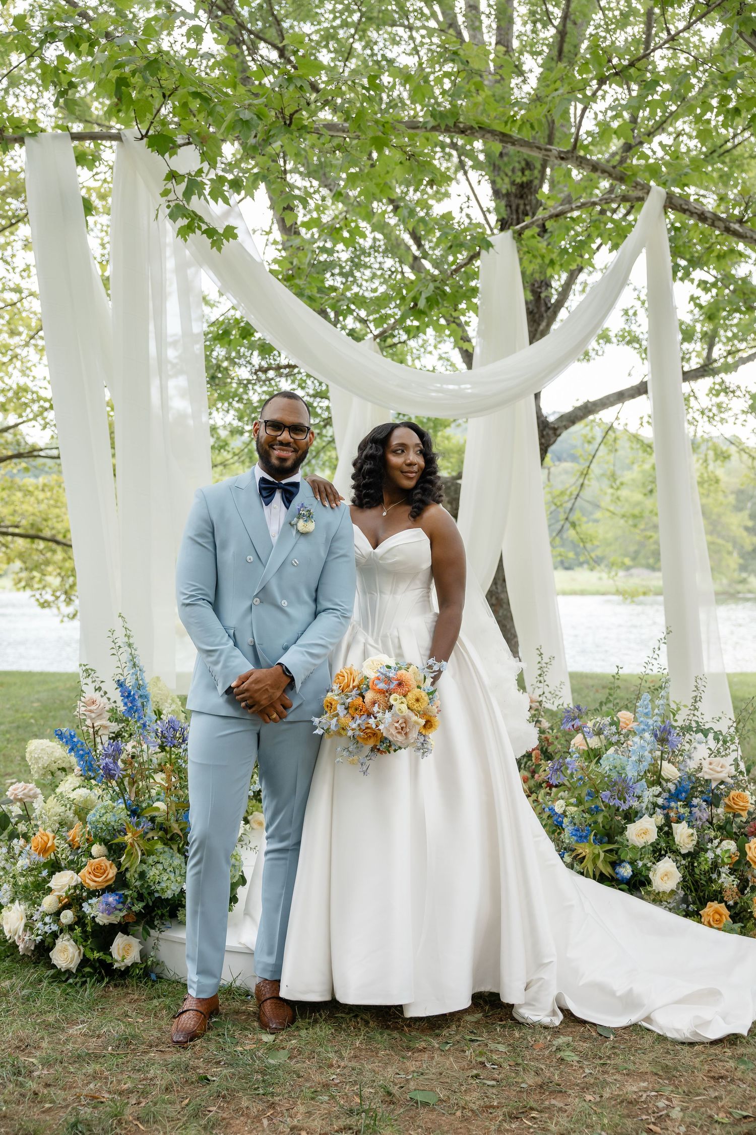 Bride and groom in light blue suit pose under white draped arch with colorful florals.
